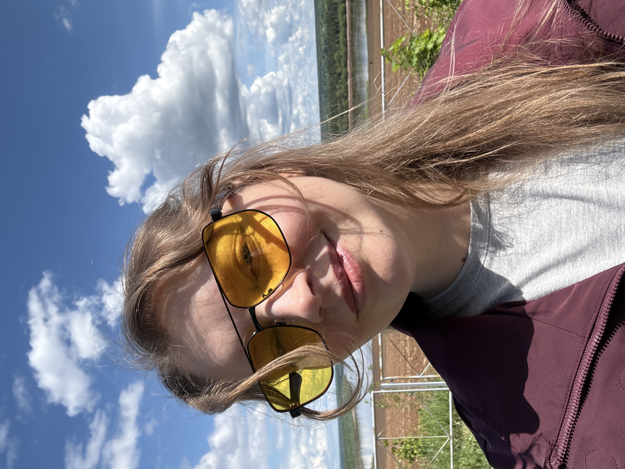 A young adult woman poses for a selfie outdoors. She is wearing yellow-tinted sunglasses, a grey shirt, and a burgundy jacket. The background features a blue sky with puffy white clouds, a field, and a wire fence. The location is seemingly rural and does not immediately resemble a city location. There is no evidence to indicate the location as Sludka, Russia.
