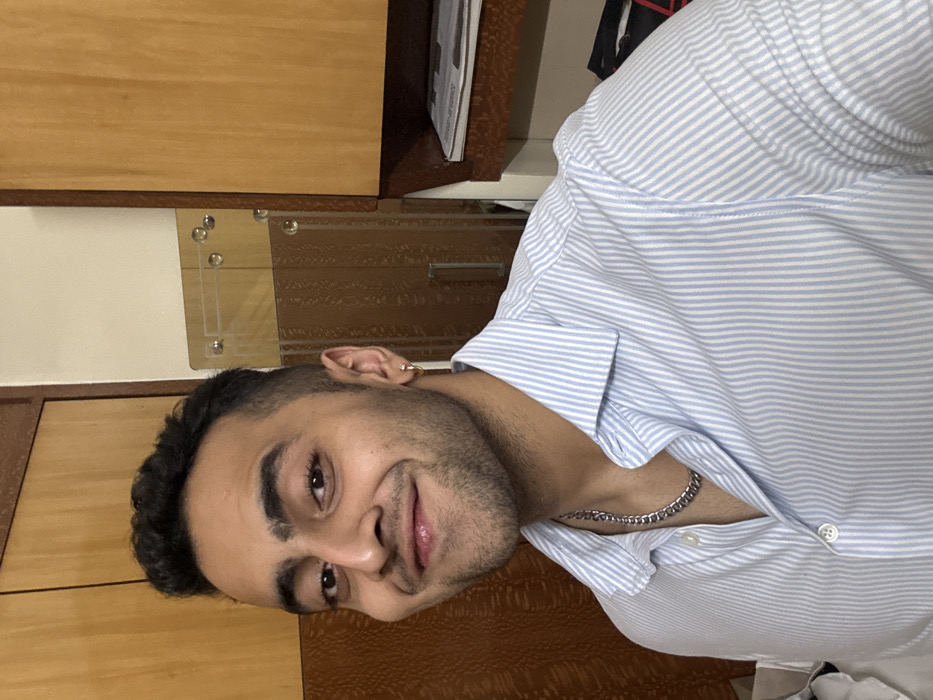A man with dark hair and a silver chain poses for the camera. He's wearing a light blue and white striped shirt. He has a small hoop earring. The background consists of wooden panels and drawers. The overall composition has a slight tilt.
