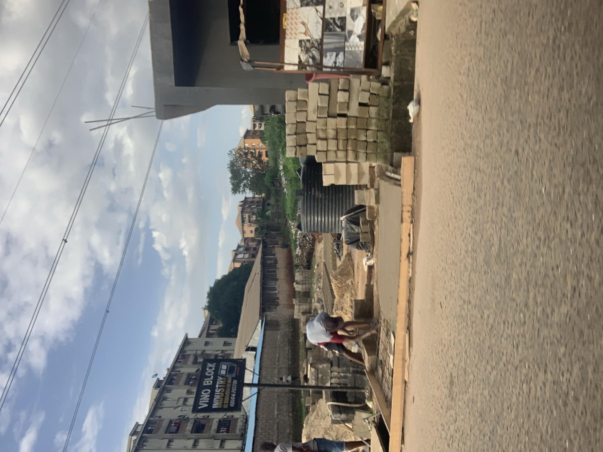 The image depicts a construction site in Nkpor, Nigeria. In the foreground, a man is kneeling and working on a section of concrete with metal reinforcement. Behind him, stacks of concrete blocks and sand are visible, along with a large black water tank. To the left, a sign for "VINO BLOCK INDUSTRY" is mounted on a pole, with a building featuring multiple balconies in the background. Further back, several residential buildings with orange and beige facades are seen amidst greenery. The sky above is partly cloudy. The overall scene suggests ongoing construction and industrial activity in a semi-urban environment.