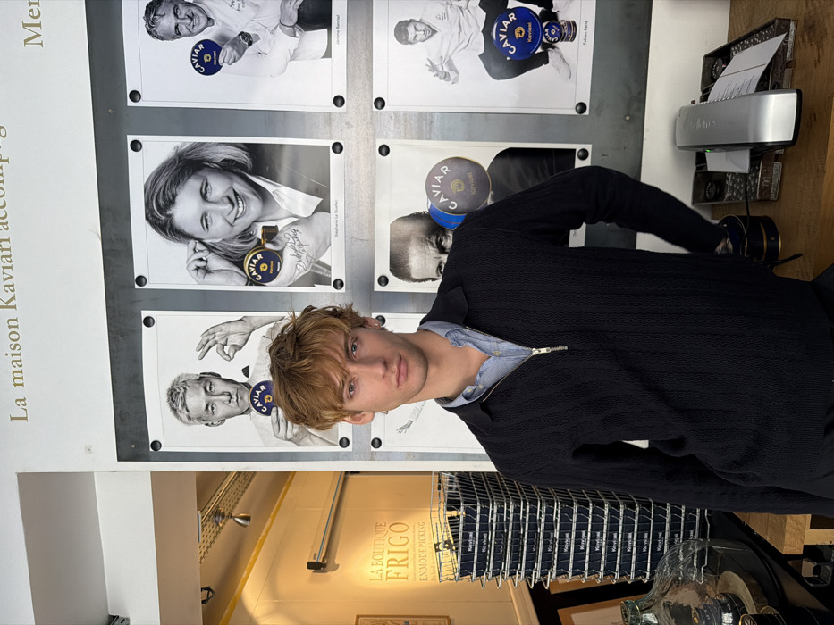 The image depicts the interior of La Maison Kaviari boutique in Paris, France, likely located in the 3rd arrondissement. A young, light-skinned male with tousled light brown hair and a neutral expression stands centrally, looking directly at the camera. He wears a dark navy textured quarter-zip pullover over a light blue collared shirt. To his left, a chrome wire basket contains a vertical stack of approximately 14 dark blue "Kaviari" branded boxes. Above and behind the basket, a white sign with orange text reads "LA BOUTIQUE FRIGO" and "EN MODE PICKING". To the male's right, on a light-colored wooden counter, a silver document shredder contains inserted paper. The background features a wall display with six framed black and white portraits of individuals, each holding a blue and gold "CAVIAR Kaviari" tin. Above these portraits, French text is visible, partially obscured, beginning with "La maison Kaviari".