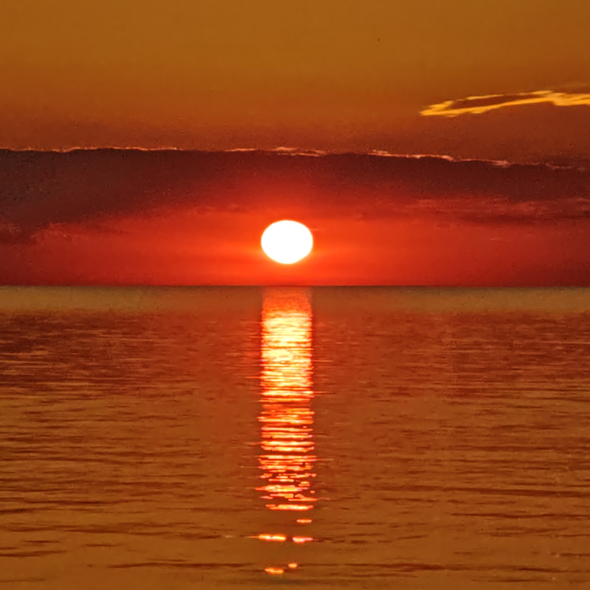 The image depicts a sunset over a body of water in an unknown location. A bright, circular, white-yellow sun is positioned low on the horizon, slightly above the vertical center of the frame. The sky above the sun is a gradient of orange, transitioning from a lighter orange-yellow at the top to a deeper reddish-orange closer to the horizon. A prominent, dark, reddish-brown cloud band stretches horizontally across the sky, obscuring part of the horizon line and positioned beneath the sun. A thin, elongated, yellowish cloud formation is visible in the upper right quadrant of the sky. The horizon line is clearly defined, separating the sky from the water. The water surface is calm, exhibiting an orange-brown hue that reflects the sky's colors. A distinct, bright, vertical column of shimmering light, reflecting the sun, extends directly downwards from the sun onto the water, gradually diffusing and widening towards the bottom of the image. The water surface shows subtle textural variations indicative of small ripples. No individuals or man-made structures are discernible in the scene.