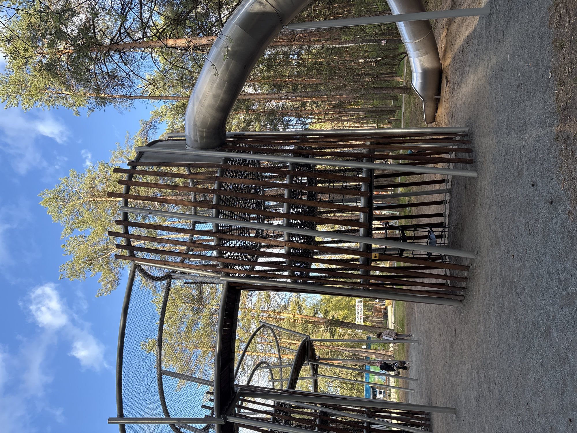 The image shows a modern playground structure located in a park. The structure consists of metal frames, wooden slats, and a tube slide. Parts of the structure are enclosed with netting. Trees are visible in the background. Two people are visible in the distance. The photo appears to have been taken outdoors on a clear day. The location is in Perm, Russia.