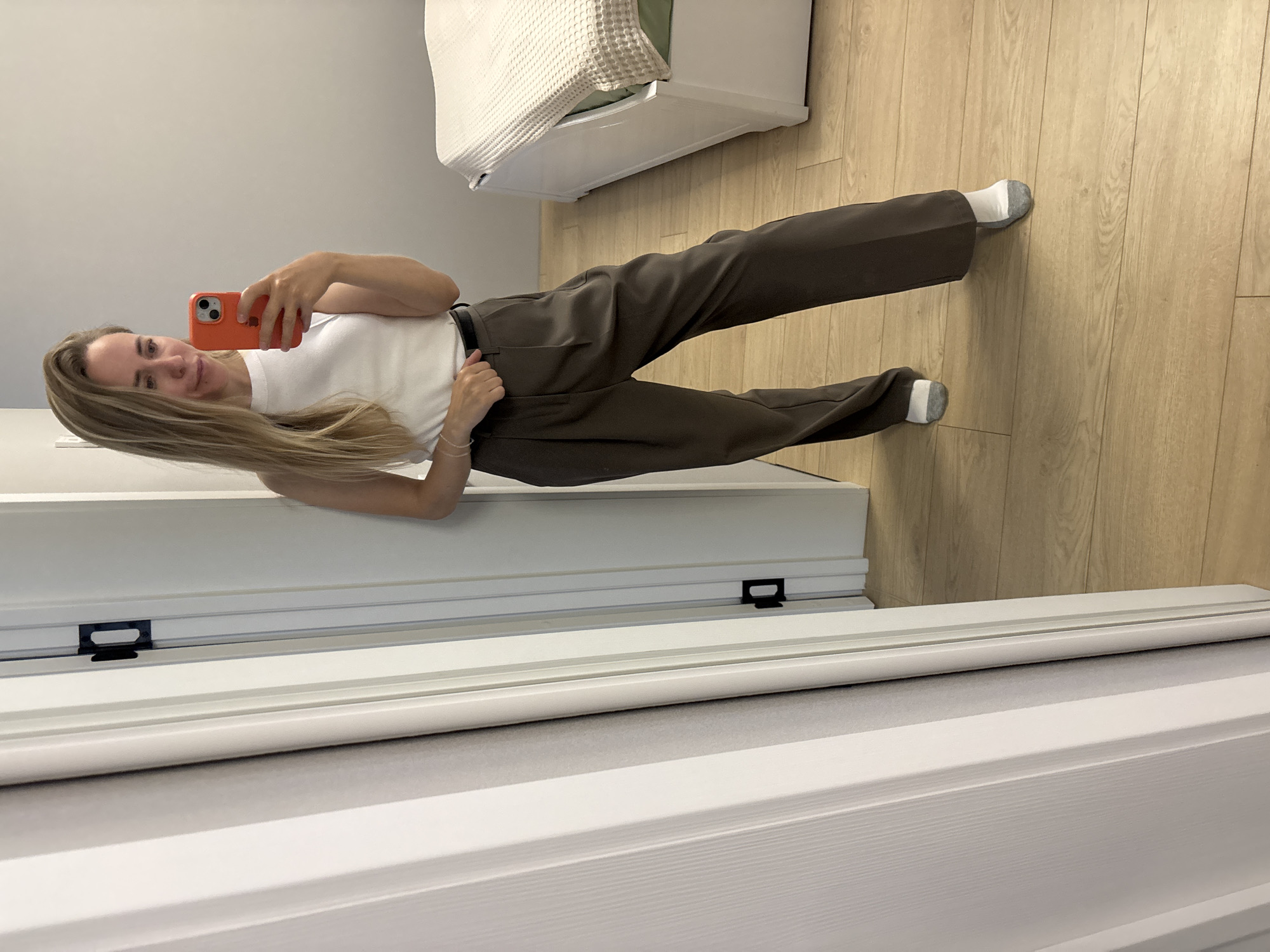 The image depicts a woman in a white tank top and dark brown trousers, standing indoors and taking a mirror selfie. She is holding an orange smartphone. The room appears to be a bedroom, with a bed visible on the right side of the frame, covered with a light-colored textured throw. The floor is made of light-colored wood planks. A white door frame and a portion of a door are visible on the left. The scene does not provide any specific indicators that would allow for its identification as being located in Chaikovsky, Russia.