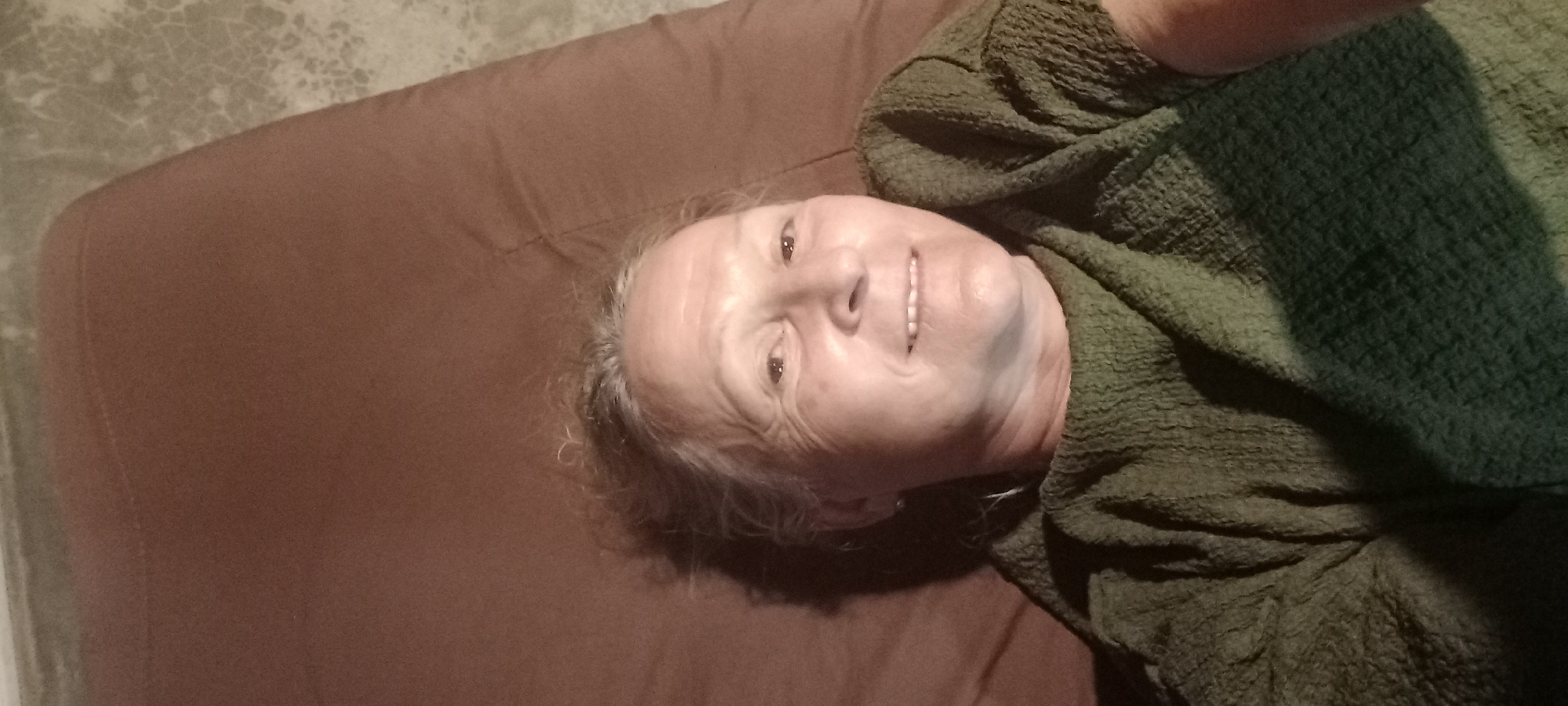 The image shows an elderly woman lying on a brown cushion or mattress. She is wearing a green, textured shawl or sweater. The background includes a textured, light gray surface, possibly a floor or wall. The image's location within the city of Ban Tak Daet, Thailand, cannot be determined from the visual information provided.