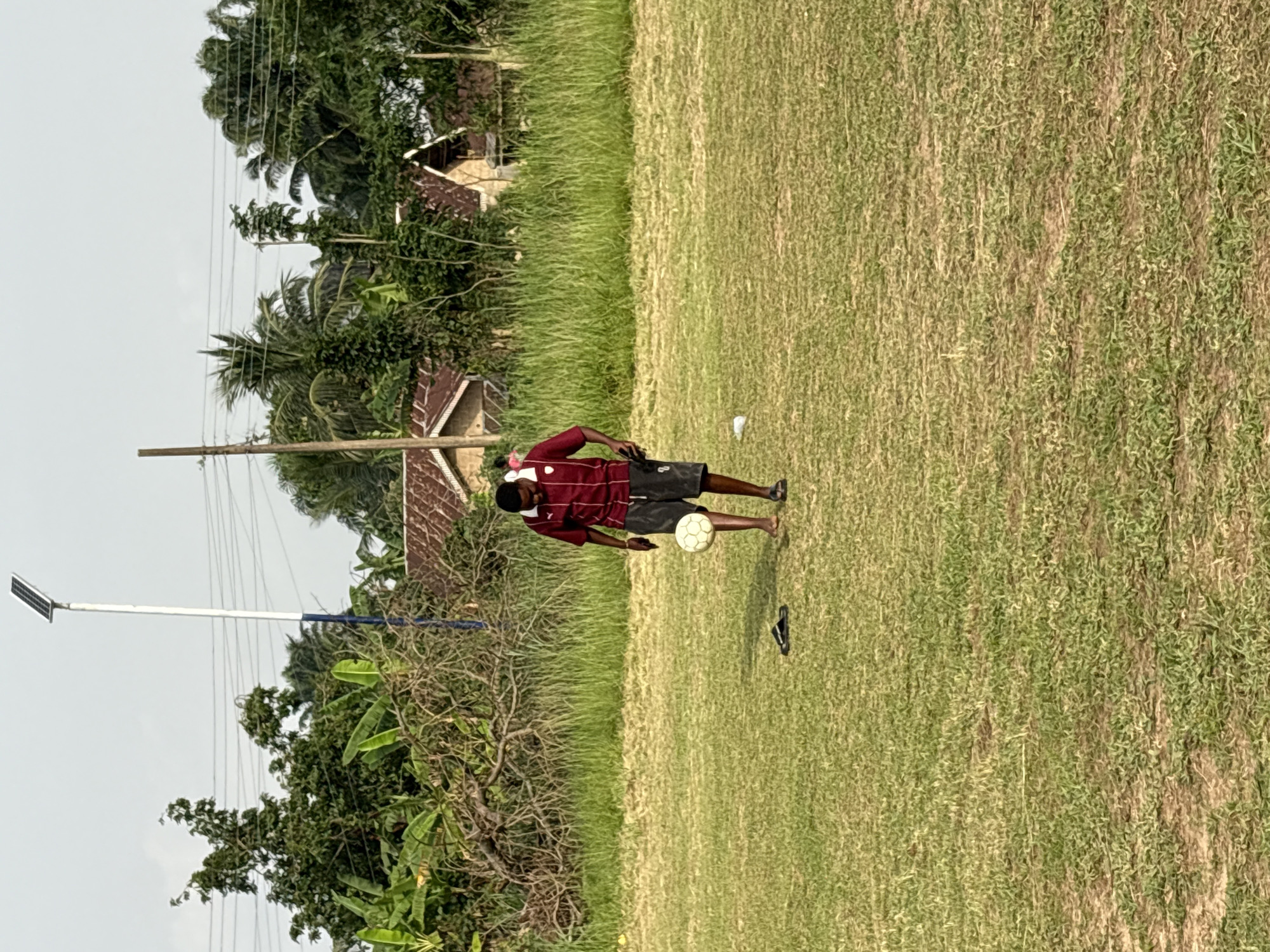 The image depicts a man standing on a grassy field in Ikot Ekpene, Nigeria. He is wearing a maroon and white striped shirt, dark shorts, and sandals. He is balancing a soccer ball on his right leg. A single sandal lies on the ground near the ball. The field has a house with a rusted roof in the background, behind trees and power lines. A solar street light pole is also visible on the left side of the image.