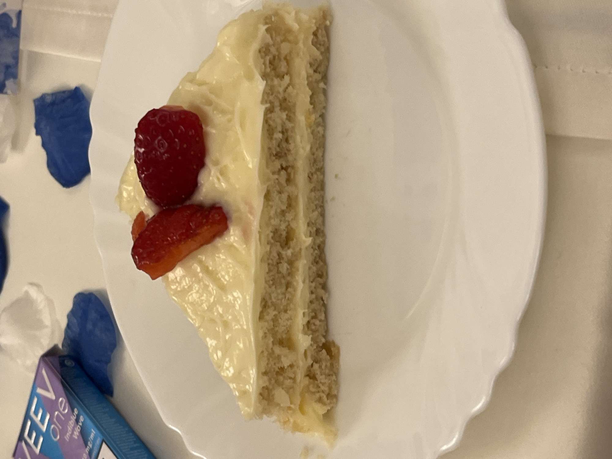 This scene, located in the city of Sofia, Bulgaria, depicts a triangular slice of cake centered on a white, scalloped-edge plate. The cake consists of two distinct layers of light-brown, textured sponge separated by a thin stratum of off-white cream or frosting. A thick, uneven layer of similar off-white cream or frosting covers the entire top surface of the cake. Garnishing the cake are two red fruit segments: one appears to be a whole, small berry (likely a strawberry or raspberry) with visible seed textures, while the other is a lengthwise cut section of a larger red fruit, possibly a strawberry. To the upper left of the plate, partially visible, is a VEEV ONE vaping product package, predominantly blue and purple, displaying text that includes "Indiblue Wave" and "18 mg/ml". Scattered around the background, particularly near the VEEV package, are several crumpled pieces of material, some blue and some white. The plate rests on a light-colored, subtly textured surface, likely a tablecloth or placemat. The lighting is soft and even, consistent with an indoor environment.