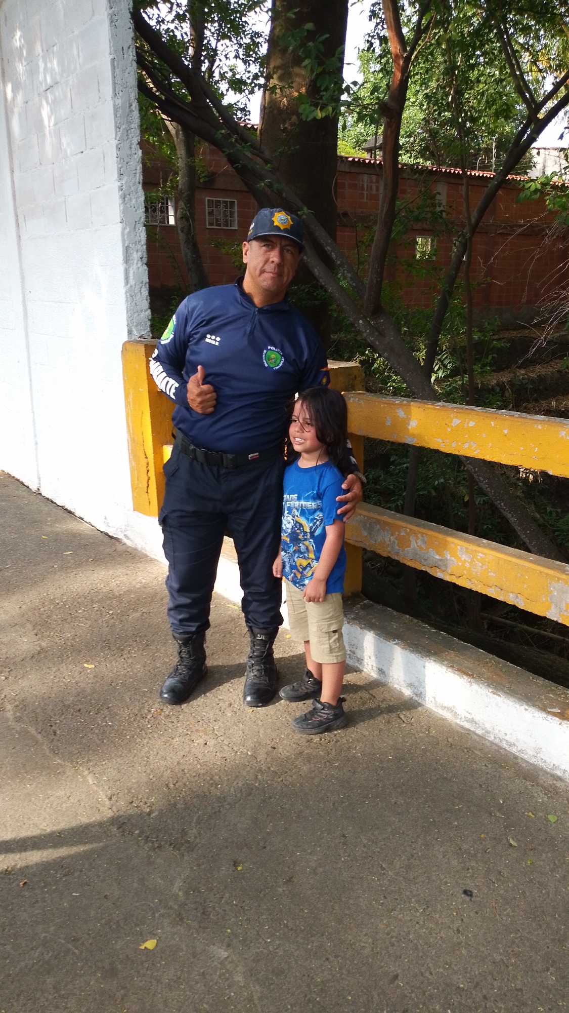 The image features an outdoor scene in Tinaquillo, Venezuela, with a man in uniform and a young child posing on a paved pathway. The man, likely a police officer, wears a dark blue long-sleeved uniform top with a half-zip, dark blue tactical pants, and black combat boots. He has a dark blue baseball cap adorned with a yellow and blue emblem, which includes the word "POLICIA" and "TRANSITO" (Traffic Police). His uniform top also displays "COMBATE" on the left sleeve and a circular green and yellow patch with "POLICIA TRANSITO" on the right chest. He is giving a thumbs-up with his right hand and has his left arm around the child.
The child, a young boy with medium-length dark hair, is wearing a bright blue t-shirt featuring a graphic of Transformers robots and the word "TRANSFORMERS" prominently displayed. He also wears light brown cargo shorts and black sneakers. The child is smiling widely, looking slightly to his left, and appears happy.
They are standing on a grey concrete surface with scattered leaves. To their right is a sturdy railing painted yellow on top and white on the bottom, suggesting a bridge or elevated walkway. The background is lush with mature trees, and behind the foliage, red brick buildings with windows can be seen. The bright sunlight casts distinct shadows on the ground, indicating it is daytime with clear weather. The overall impression is a warm, candid moment captured between the two individuals.
