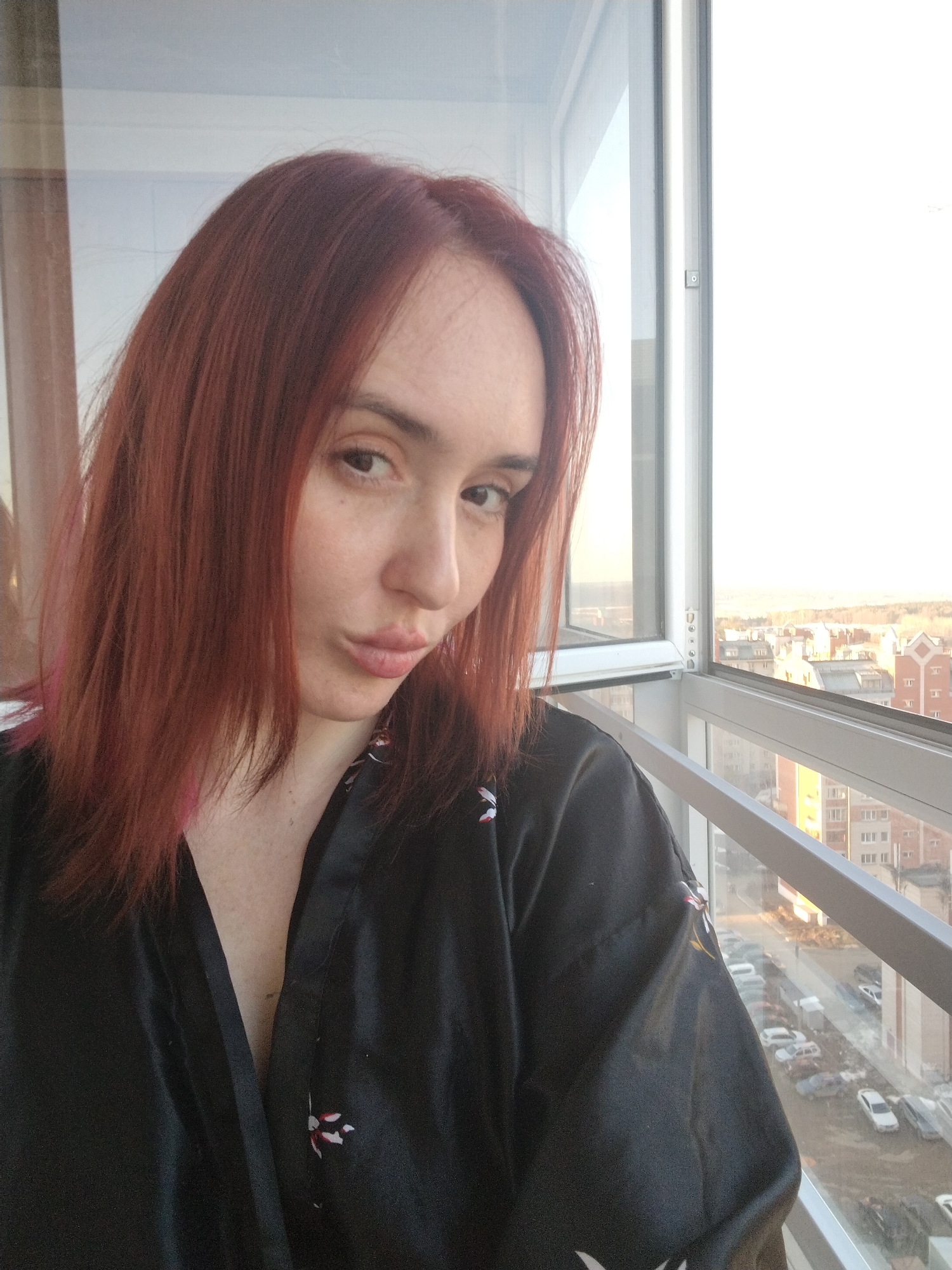 A woman with shoulder-length auburn hair is posing for a selfie in front of a window in Izhevsk, Russia. She is wearing a black satin robe with a floral pattern. The window overlooks a cityscape with apartment buildings and parked cars. The time of day appears to be late afternoon or early evening, given the light.