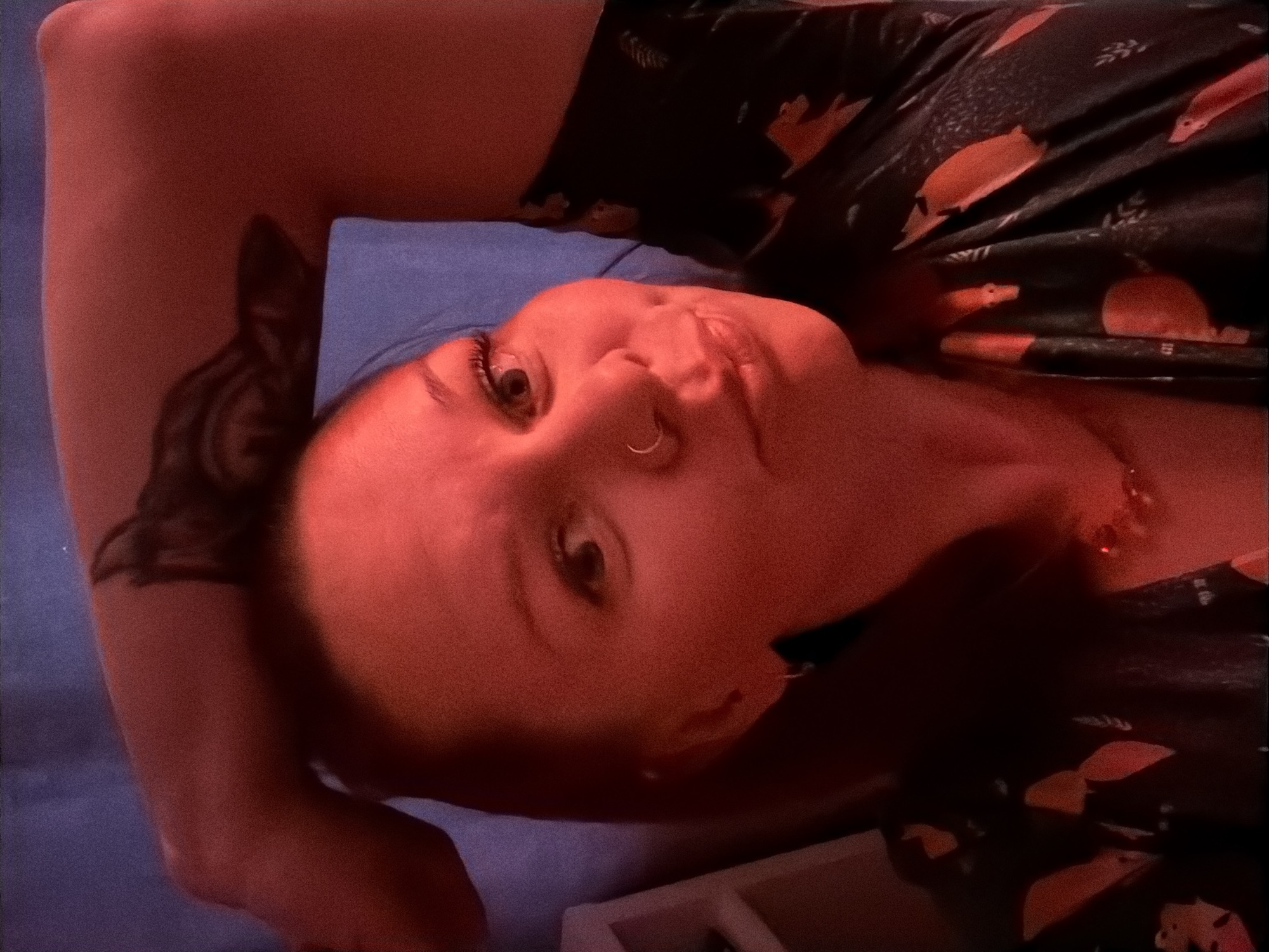 A woman with fair skin, a nose ring, and a tattoo on her upper left arm is lying on a blue surface. She is wearing a dark shirt with orange and white patterns. The image is predominantly lit with a red hue. There is no visible indication of the location being Chaikovsky, Russia.