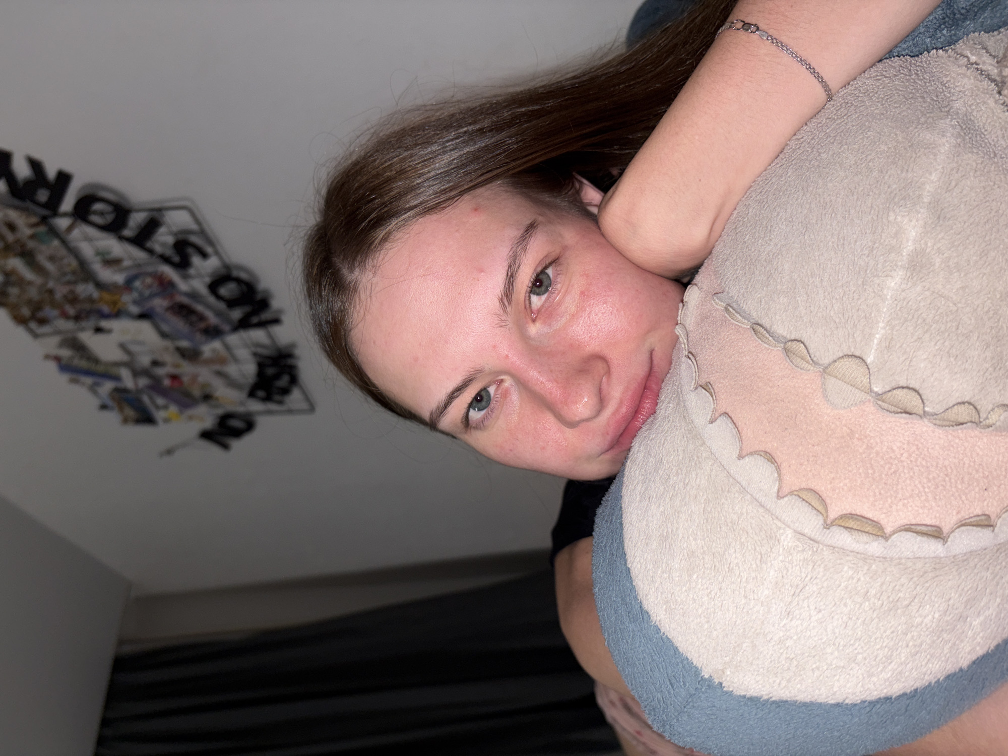 An indoor, eye-level, medium shot shows a young woman resting her head on a soft, multi-colored pillow in what appears to be a bedroom. The woman has long, brown hair, fair skin with some redness and blemishes, and blue eyes. She is looking directly at the camera with a relaxed expression. Her right arm is bent, supporting her head, and she is wearing a dark t-shirt. A delicate silver bracelet is visible on her wrist. Behind her, a wall is decorated with a wire frame collage of photos and letters that spell out "NO RISK NO STORY." A dark curtain is visible on the left side of the frame. The lighting is dim, suggesting it is nighttime or the room is dimly lit. There is no identifiable information about the location being in the city of Perm, Russia.