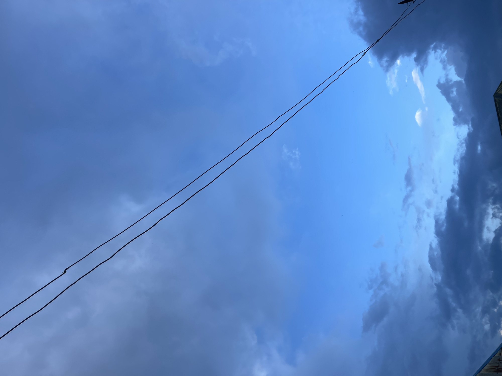 The image depicts a dramatic sky over what appears to be a semi-urban or rural environment in Rafin Par, Nigeria. The sky is a deep blue, transitioning from lighter hues near the horizon to a darker shade overhead. Heavy, dark clouds dominate the lower portions of the frame, suggesting an impending storm or recent rainfall. These clouds are contrasted by scattered patches of brighter, white clouds higher in the sky.
Stretched diagonally across the frame are two dark power lines, their thin profiles stark against the turbulent sky. One of the lines appears to have a small, tangled section, possibly indicating damage or an obstruction. The upper left and lower right corners of the image show glimpses of structures, likely buildings with angled roofs, partially obscured by the low-lying clouds and darkness. No people are visible in the image. The overall impression is one of impending weather change and a sense of the raw power of nature in this Nigerian setting.