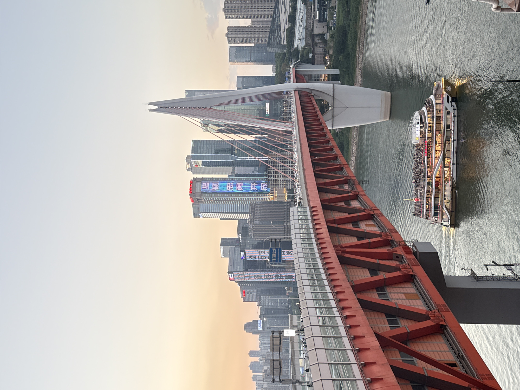 The image captures a view of the Hongyadong Bridge in Chongqing, Yuzhong District, China, during what appears to be sunset. The bridge, characterized by its distinctive red superstructure and white pylon, spans a wide body of water. On the bridge, traffic is visible, indicating it is an active thoroughfare. Below the bridge, a large, multi-deck passenger boat, illuminated with warm lights and carrying numerous passengers, is moving through the water, leaving a wake. In the background, a dense cityscape rises, dominated by modern skyscrapers. The sky above transitions from a soft orange hue near the horizon to a pale blue, suggesting the twilight hours. The perspective is from a slightly elevated position, looking towards the city and the bridge.