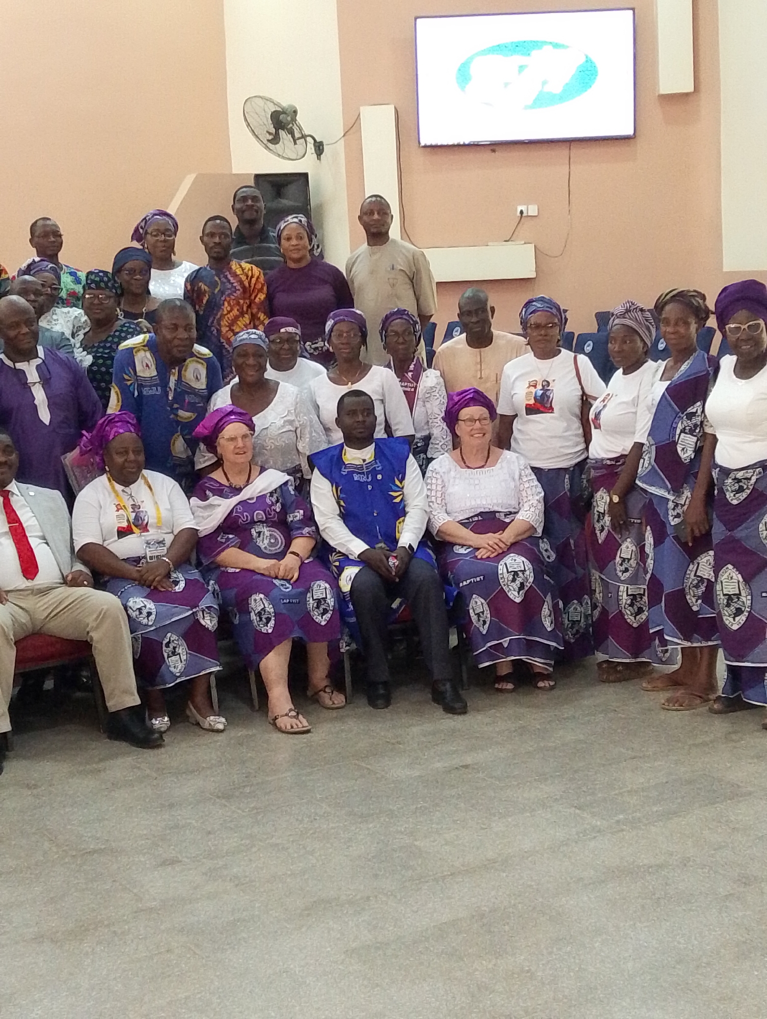 This full-shot photograph captures a large group of approximately 40 adults, predominantly men and women, posing for a group portrait indoors in Kubwa, Nigeria. The subjects are arranged in multiple rows, with some seated in the foreground and others standing behind them.
Many of the women are dressed in coordinated attire, consisting of purple and white patterned skirts or wraps, often paired with white blouses or t-shirts, and matching purple head wraps. The word "BAPTIST" is clearly visible in white text on the patterned fabric of several women's skirts, suggesting a religious affiliation. Some men are also wearing coordinated blue and white patterned vests or shirts, featuring letters such as "B.J.U.". Other individuals wear a mix of traditional African attire and more casual Western clothing. Many attendees wear glasses, and some have lanyards or badges, though their text is illegible.
The setting appears to be an indoor hall or meeting room, with plain light-colored (peach/beige) walls and a light grey floor. In the background, a large flat-screen television is mounted on the wall, displaying a logo which is not clearly identifiable but shows a symbol with some light-colored text on a white background. A wall-mounted fan is visible on the right side of the image, alongside a dark speaker or piece of equipment. The subjects generally display pleasant and formal expressions, looking towards the camera. The matching attire and visible "Baptist" text strongly suggest this gathering is a conference, convention, or special event related to a Baptist church or organization.