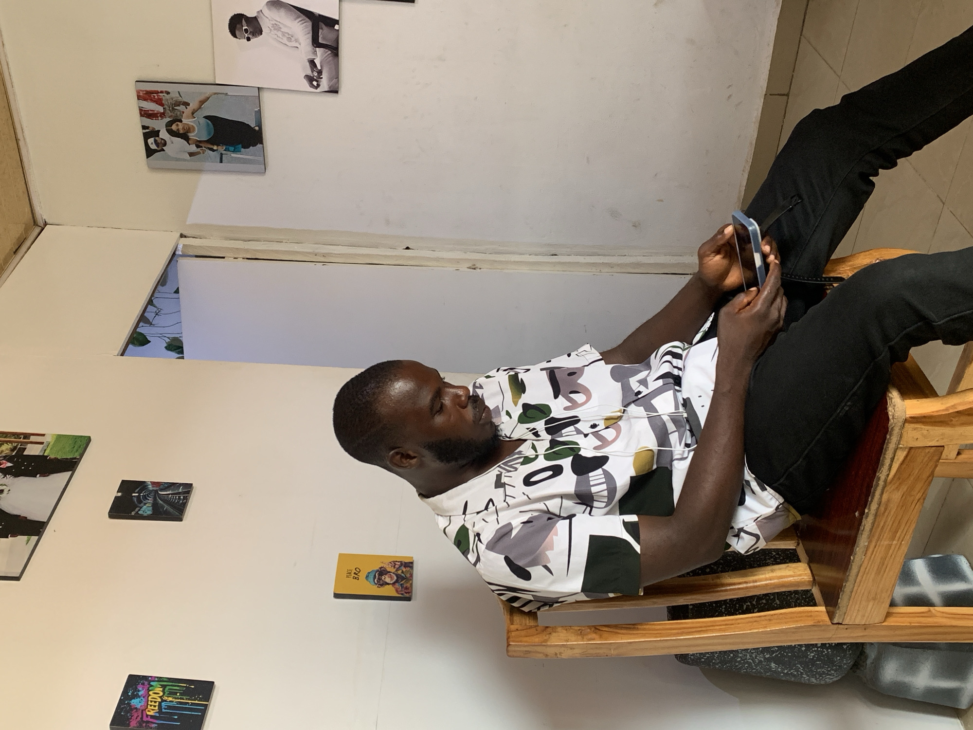 A person is seated in a wooden chair, looking at a mobile phone. The person is wearing black pants and a patterned short-sleeved shirt. The location appears to be indoors, with various framed images on the wall. The location is not specified in the image.