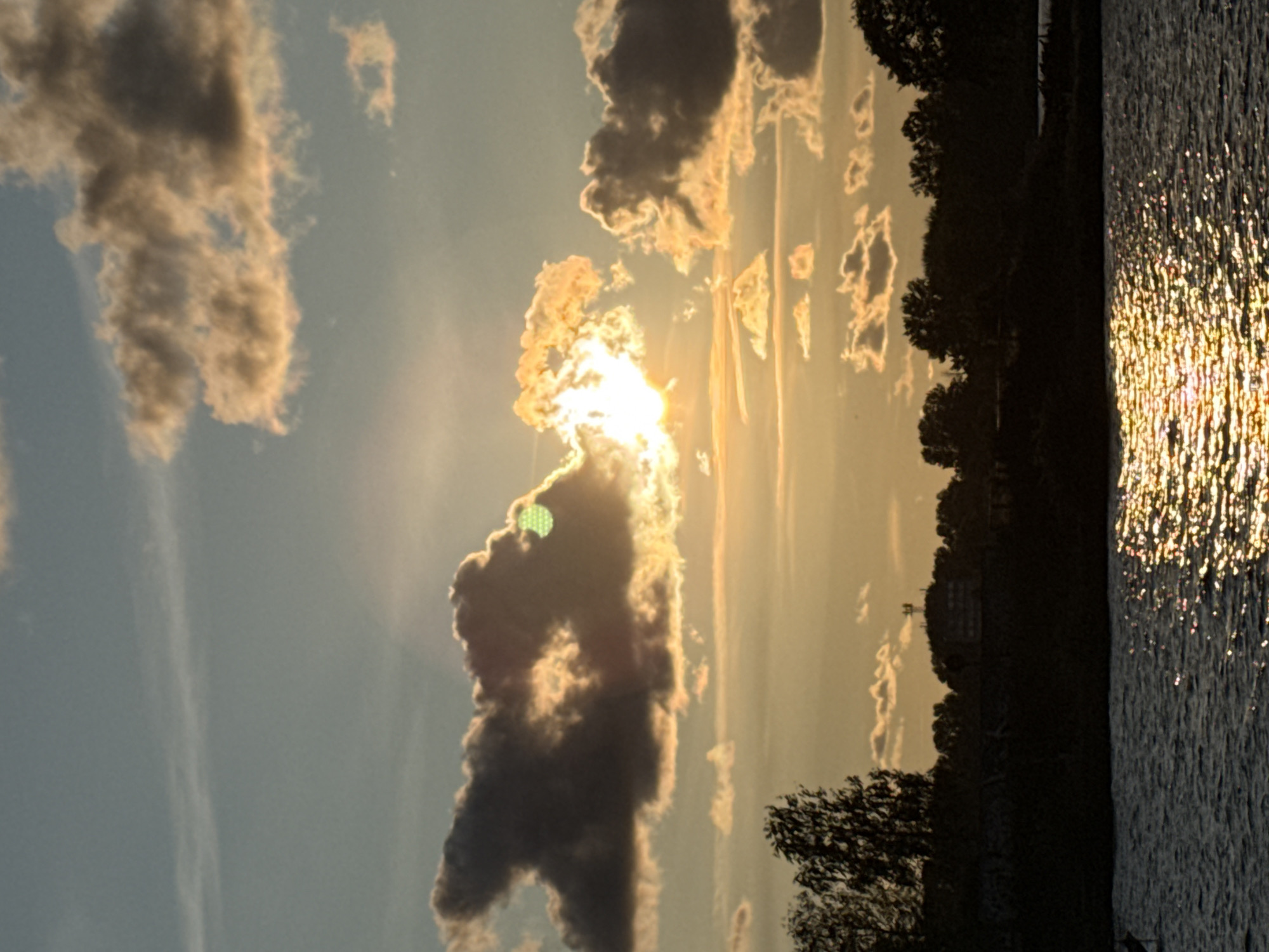 This is a photograph of a sunset over a body of water, likely a river or lake, with a silhouetted shoreline. The sky is filled with a mix of light blue and orange hues, with scattered clouds catching the golden light of the setting sun. The sun itself is partially obscured by dark clouds, creating a dramatic effect with rays of light emanating from behind them. The water in the foreground reflects the warm colors of the sky, creating a shimmering path of light. The shoreline is a dark silhouette of trees and some man-made structures, including what appears to be a building with an antenna or tower. The scene evokes a sense of peace and natural beauty.
While the image depicts a sunset over water with a silhouetted shoreline, there are no specific visual cues or landmarks within the photograph that definitively place it in Riga, Latvia. The general landscape could be found in many locations.