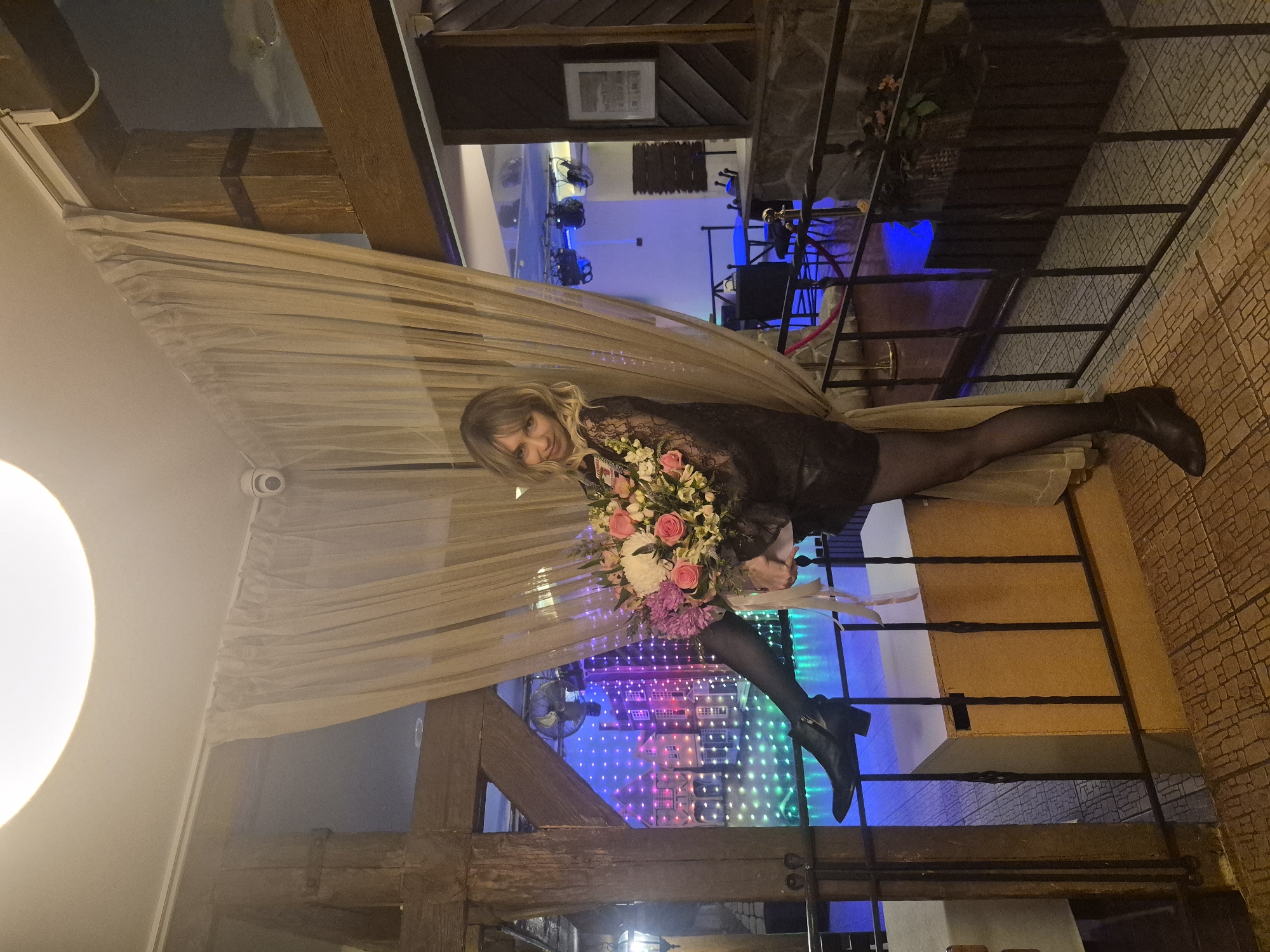 A woman is sitting on a ledge next to a railing in a room with a high ceiling. She is wearing a dark-colored dress, sheer black stockings, and black boots. She is holding a bouquet of flowers. The room is decorated with drapes and wooden beams. In the background, there is a view of a room with colorful lights. The location is in Chaikovsky, Russia.