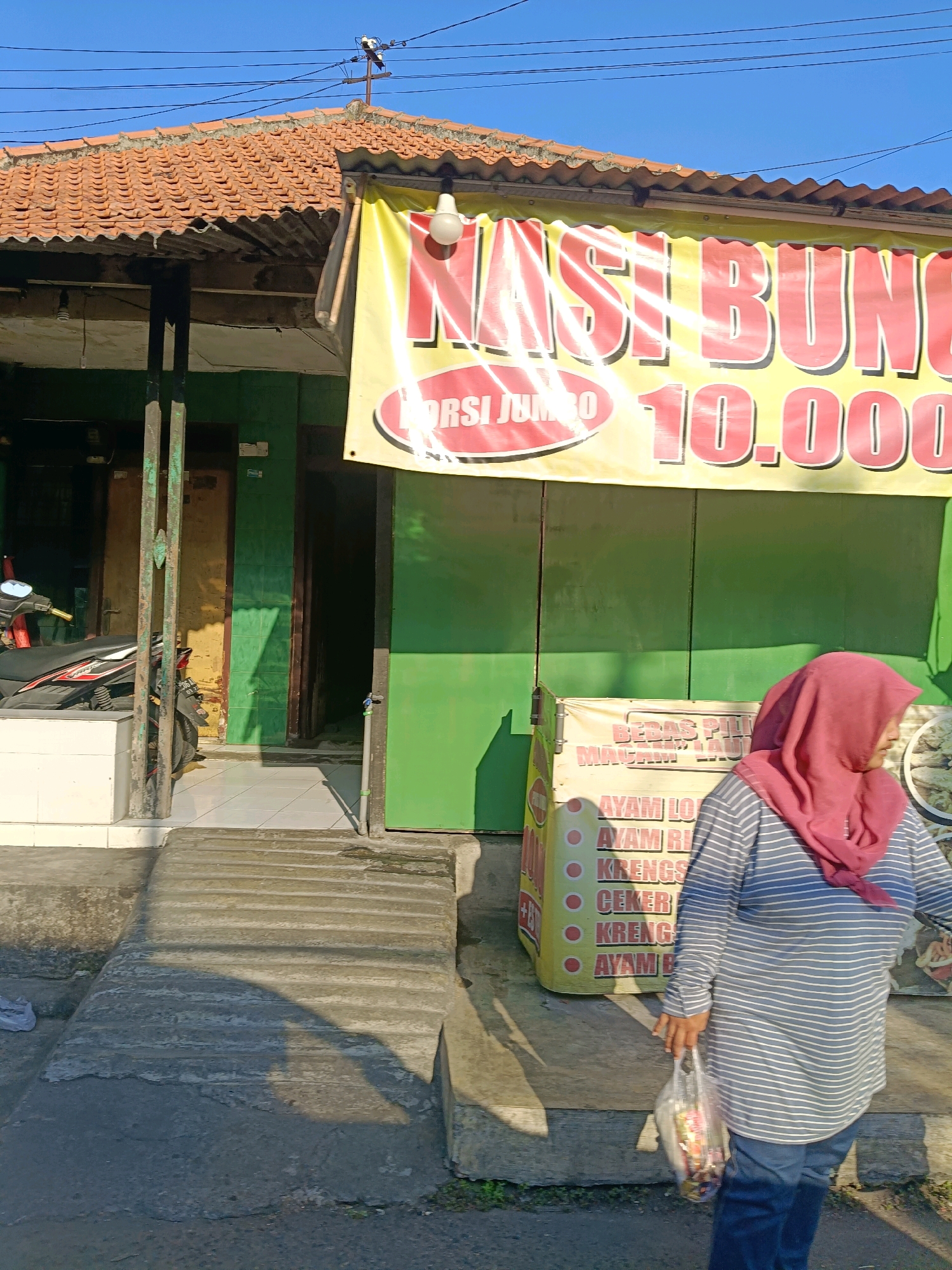 The image shows a street view in Surabaya, Indonesia, with a small building that appears to be a food stall or small shop. A large yellow banner with red lettering advertising "NASI BUNGKUS JUMBO 10.000" (Jumbo Rice Package 10,000) is prominently displayed across the front of the building. The building has a tiled roof and green walls. In front of the building, a person wearing a pink hijab and a striped long-sleeved shirt is walking away from the camera, carrying a clear plastic bag filled with items. To the left of the entrance, a motorcycle is parked. The ground in front of the building is a concrete ramp with textured steps. The overall environment suggests a sunny day in a bustling urban area.