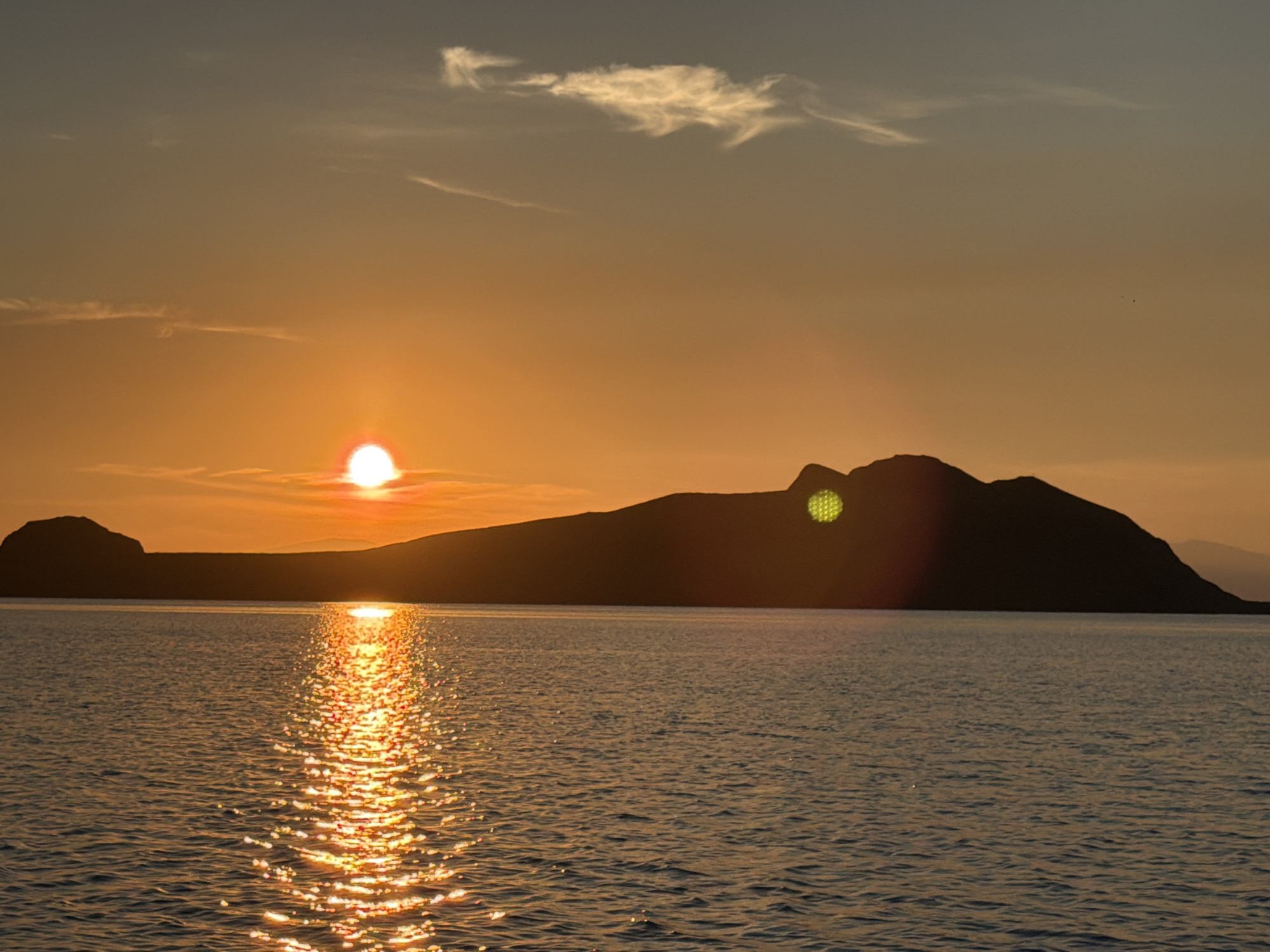 The image shows a sunset over a body of water, with a silhouette of a mountainous island in the background. The sun is a bright orange orb low in the sky, casting a shimmering reflection on the water. The sky is a gradient of orange and gray, with wispy clouds. The water is calm, with small ripples. The island is dark and rugged, with a few indistinct shapes that could be smaller islands or peninsulas. There are no people or man-made objects visible in the image. The location is described as Unknown, Russia, but this is likely a placeholder as no specific landmarks are visible to confirm this.