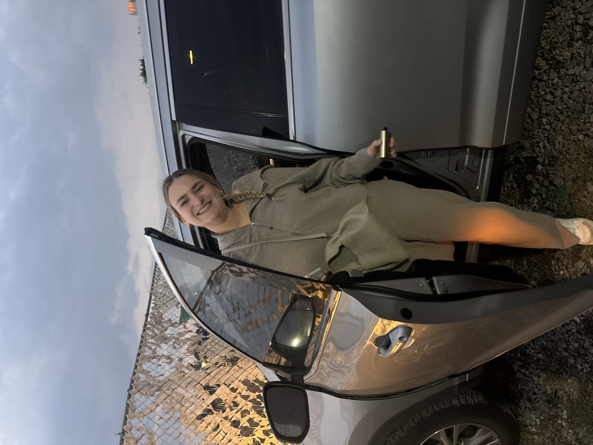 A young woman with blonde hair styled in a braid smiles from inside a silver car. She is wearing a beige jumpsuit and white sneakers. She holds a cylindrical object in her right hand. The car door is open, revealing the interior and the surrounding gravel ground. In the background, there is a cloudy sky and a fence covered with patterned material. The location is San Bernardino Tlaxcalancingo, Mexico.