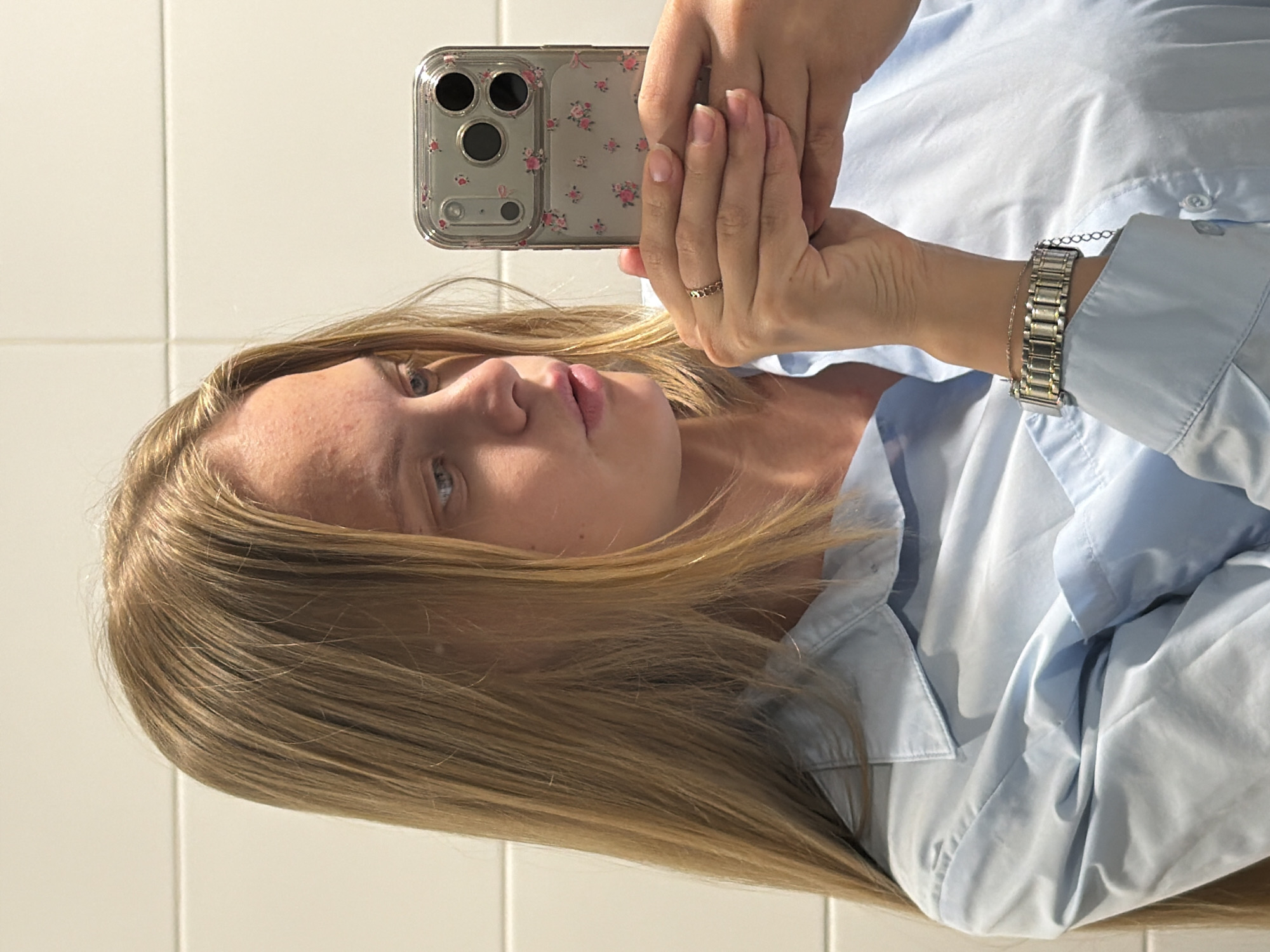 A person with blonde hair is taking a selfie with a smartphone in a clear case with a floral design. The person is wearing a light blue button-down shirt and a silver watch. The background is a white tiled wall. There is no information provided to suggest the location is Izhevsk, Russia.