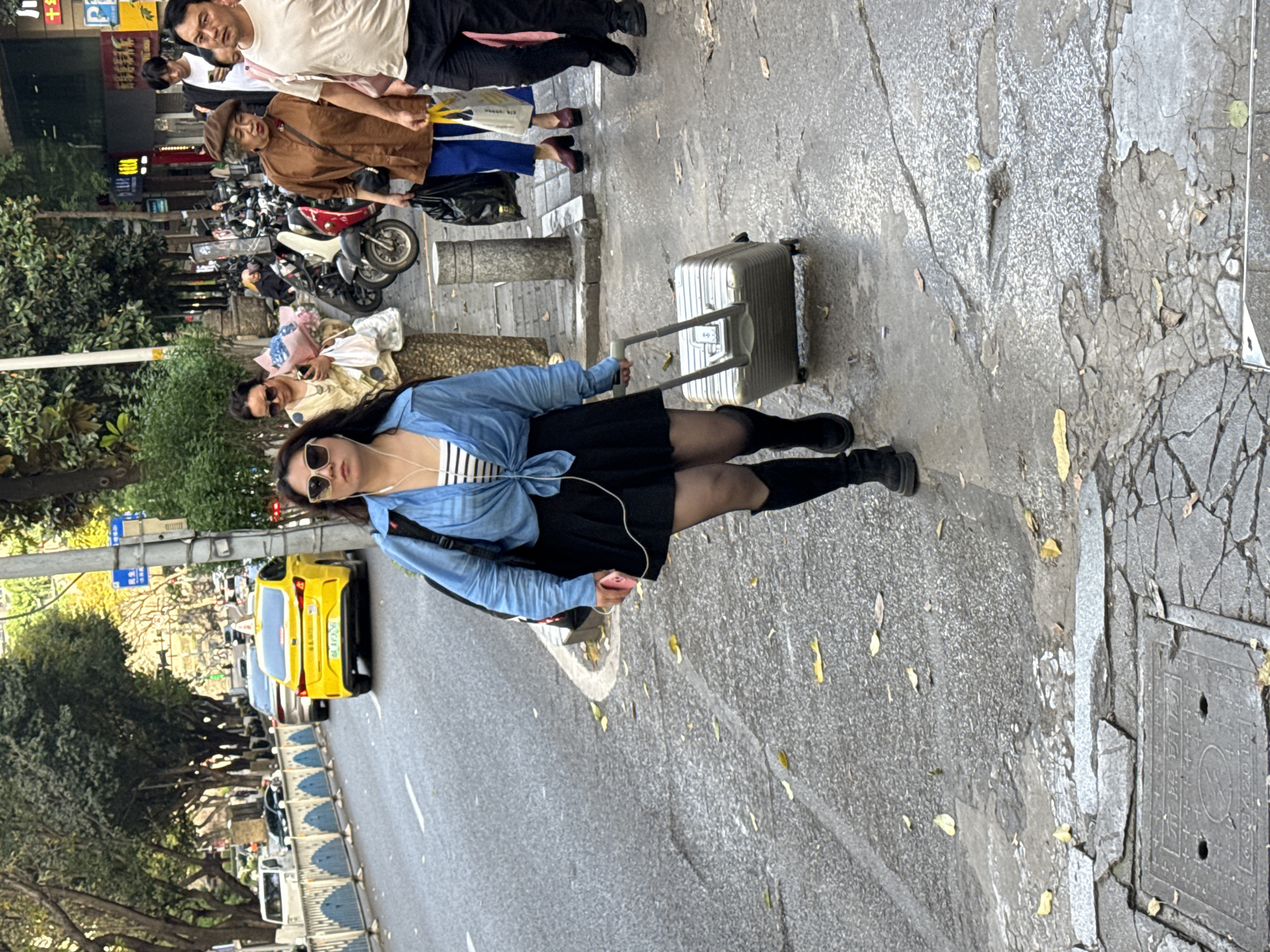 A medium, tilted outdoor shot shows a woman pulling a silver suitcase along a paved street in Yuzhong District, China. The woman, facing forward, wears sunglasses, a blue denim shirt tied at the waist, a striped top, a black skirt, black tights, and black boots. She grips the handle of the silver suitcase, which is positioned behind her on the cracked sidewalk. To the left, a yellow taxi cab is seen on a city street lined with trees and other vehicles. In the background, on the right side of the frame, there is sidewalk damage and a metal sewer cover. In the distant background, there are pedestrians.