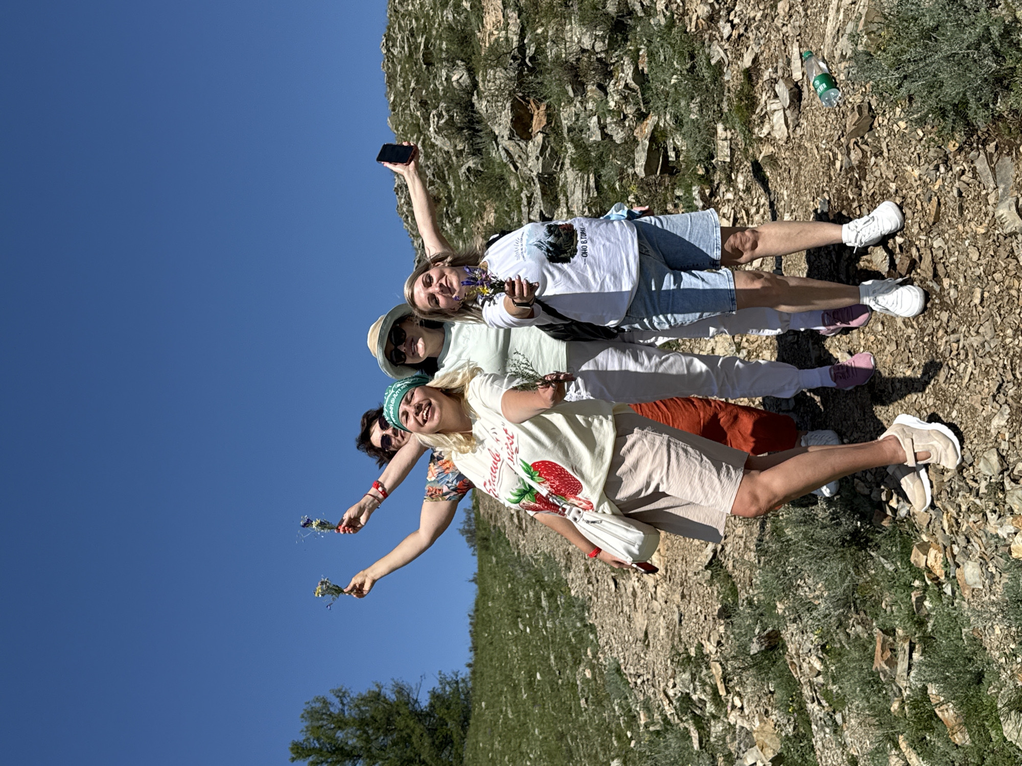 Four individuals are posing for a photograph in a mountainous, outdoor environment with a clear blue sky overhead. The scene appears to be a hiking trail in the Bichiktu-Boom Gorge, Russia, characterized by rocky terrain, sparse green vegetation, and a large tree on the left.
The individuals are standing on a dirt path. The person in the foreground, on the left, is a woman with blonde hair, wearing a white t-shirt with a strawberry graphic and the words "STRAWBERRY MIST", beige shorts, and light beige sneakers. She is holding a small bouquet of wildflowers in her right hand and a white purse on her left arm.
To her right is another woman with blonde hair, wearing a light green long-sleeved shirt, white pants, and pink sneakers. She is wearing a straw hat and sunglasses, and holding a phone in her outstretched right hand, possibly taking a selfie. She is also holding a small bouquet of wildflowers.
Behind and to the left of the first woman is a third person, whose gender is difficult to determine, wearing a colorful floral shirt and holding a small bouquet of wildflowers.
The fourth individual is partially visible behind the other three, wearing a white t-shirt with a graphic that includes text and an image of a mountain. This person is also holding a smartphone.
The overall atmosphere is casual and celebratory, with the individuals appearing to be enjoying an excursion. A plastic water bottle is visible on the ground to the right.