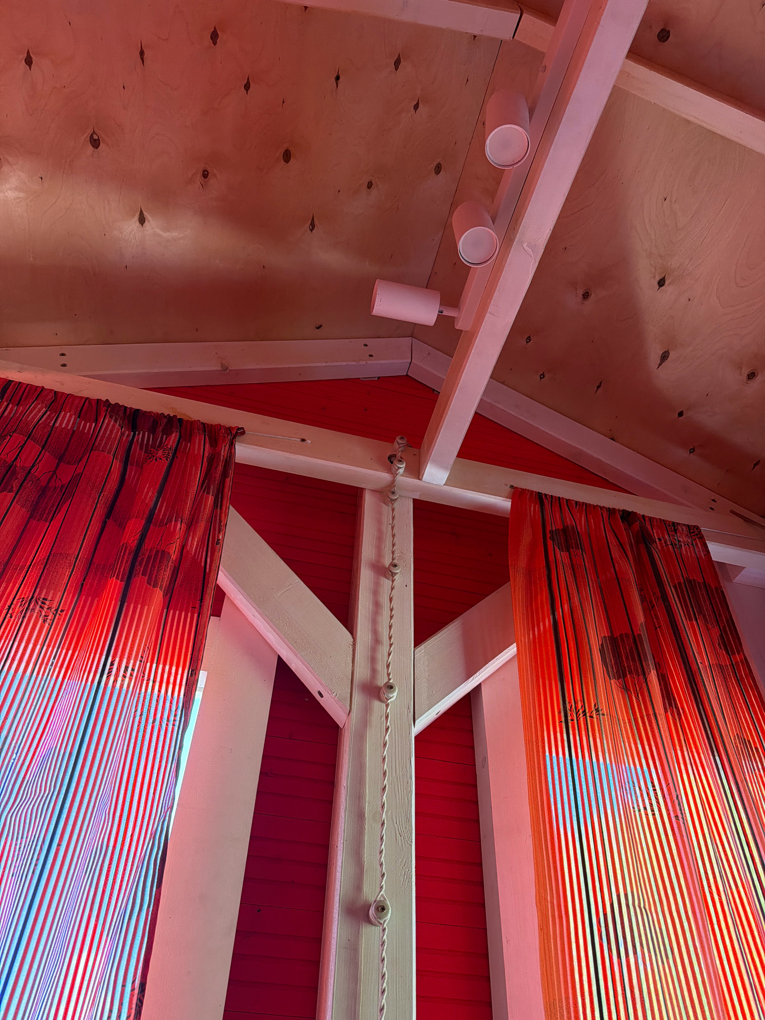 The image depicts an interior view of a structure with a sloped wooden ceiling and red-paneled walls. Two red curtains with a striped pattern and floral motifs hang on either side of a central wooden beam. The beam has a rope with ceramic insulators running along its length. Track lighting with cylindrical fixtures is mounted on the ceiling. The overall aesthetic suggests a stylized interior, possibly a room or a pavilion. The location in Stupino, Russia is not evident from the visual information provided in the image.