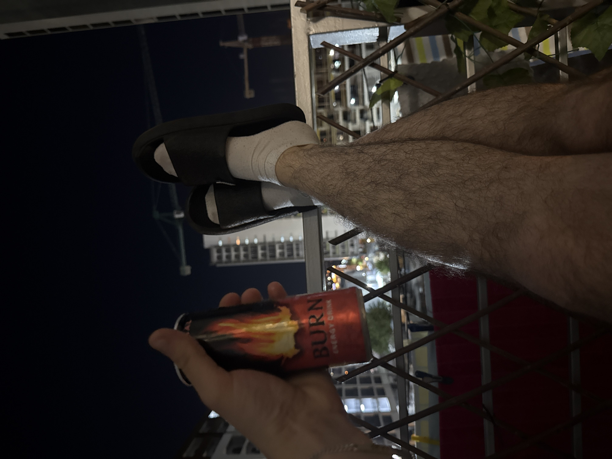 A person's hand holds a red can of "Burn Energy Drink" with flames depicted on the label. The person is wearing black slides and white socks. Their legs, covered in dark hair, are crossed and visible from the knees down. The scene appears to be on a balcony or similar outdoor space, with a metal railing and a lattice fence in the foreground. In the background, city buildings and construction cranes are visible against a dark night sky. The overall impression is that of a person relaxing outdoors at night in an urban environment.