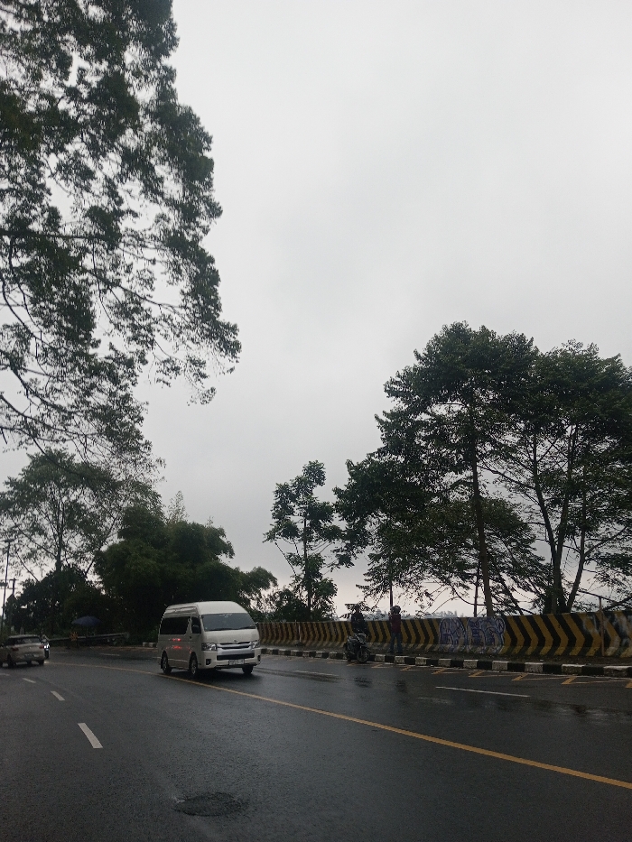 The image captures a road scene on what appears to be an overcast or rainy day, indicated by the wet asphalt and the uniformly gray sky. The perspective is from a low angle, looking slightly upwards and along the road.
The dominant feature of the sky is its muted, light gray tone, completely covered by clouds with no visible sun. Large, verdant trees frame the upper left and right portions of the image, with their dense green foliage silhouetted against the bright sky. More trees and lush greenery are visible further down along both sides of the road, suggesting a semi-forested or rural setting.
The road itself is a two-lane asphalt surface, visibly wet and reflecting light, indicating recent rainfall. A solid double yellow line runs down the center, separating the directions of traffic, and a dashed white line is present on the left side, marking the edge of a lane. A patch of standing water or a pothole is visible in the foreground on the left lane.
Vehicles are present on the road. A white passenger van, resembling a Toyota Hiace, is centrally located, driving towards the viewer on the left lane. Its headlights appear to be on, possibly due to the dim lighting conditions. Further in the distance on the left, another vehicle, likely a car, is partially visible. On the right side of the road, a concrete barrier with distinctive black and yellow chevron stripes separates the road from what appears to be a drop-off or embankment. Two individuals are standing next to this barrier; one is seated on a motorcycle parked on the shoulder, and the other is standing nearby. Some faint, illegible markings or graffiti can be seen on the barrier in this area. A small blue umbrella or canopy is faintly visible behind the trees on the far left, suggesting a potential vendor or shelter.
Overall, the image depicts a journey on a damp road, surrounded by natural elements, under a cloudy sky.