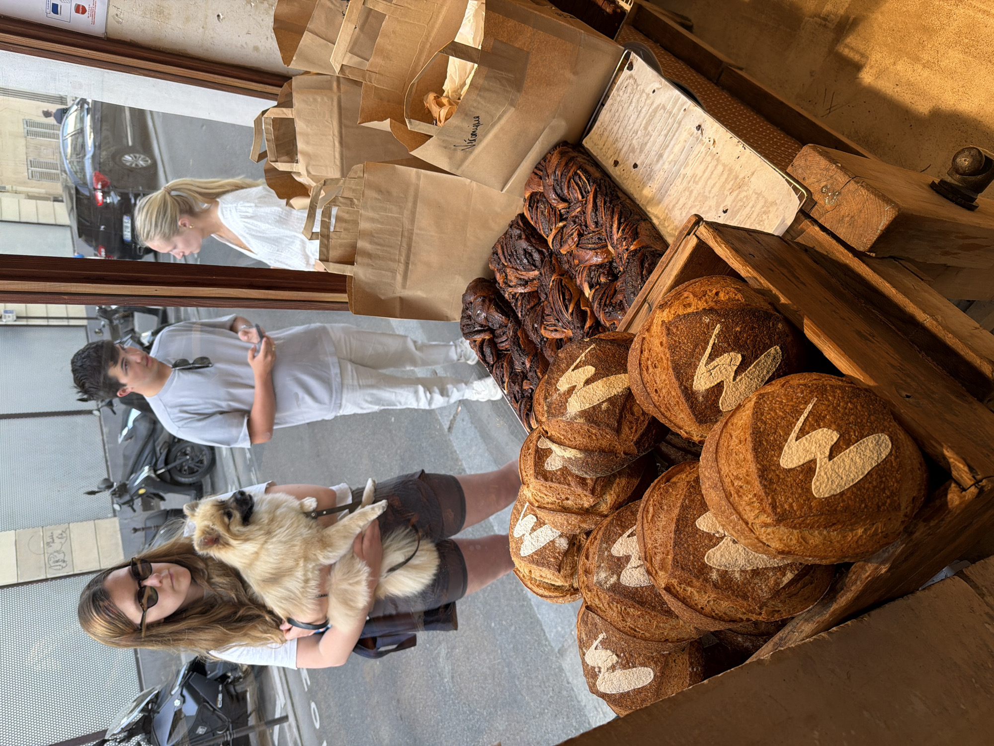The scene is captured through a window of what appears to be a bakery in Paris, France. The bakery display showcases an assortment of baked goods, including round loaves of bread marked with a white design, and a stack of braided chocolate pastries. Several brown paper bags are filled with baked goods, labeled with the bakery's name in cursive script.
The window reflects the activity outside. A woman is holding a tan dog on the sidewalk, wearing sunglasses and a casual outfit. A man, slightly out of focus, is beside her, looking towards the bakery. Behind them, a parked motorbike and a car can be seen on the Parisian street. Another woman is seen browsing inside the bakery.