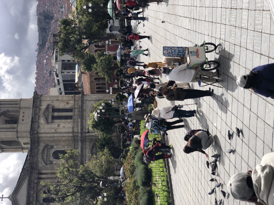 A bustling city square showcases diverse activity. A grand building with a tall tower borders the left side, shadowed by clouds. The square, paved with gray tiles, is populated with people. Many stroll, some under umbrellas, while others gather around a vendor with a cart. Near the bottom, a person kneels, surrounded by pigeons. Buildings cascade down a hill in the background under a cloudy sky.