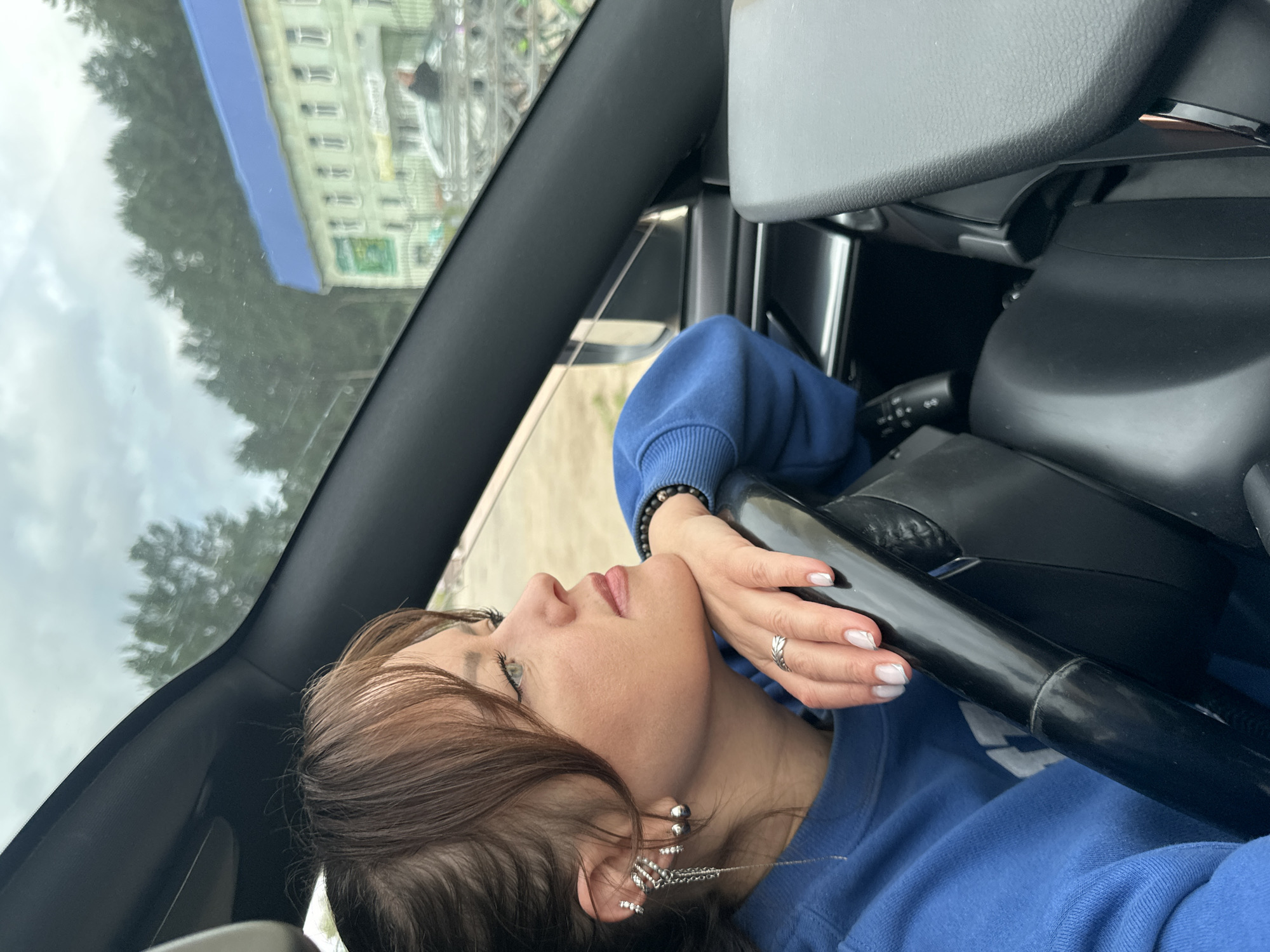 A woman with brown hair and bangs, wearing a blue sweatshirt and numerous silver earrings, is looking out the window of a car. She is resting her head on her hand, which is on the steering wheel. Her nails are painted a light color. Outside the car, a green building with a blue roof and trees are visible. The image appears to have been taken in Chaikovsky, Russia, as indicated by its geographical context.
