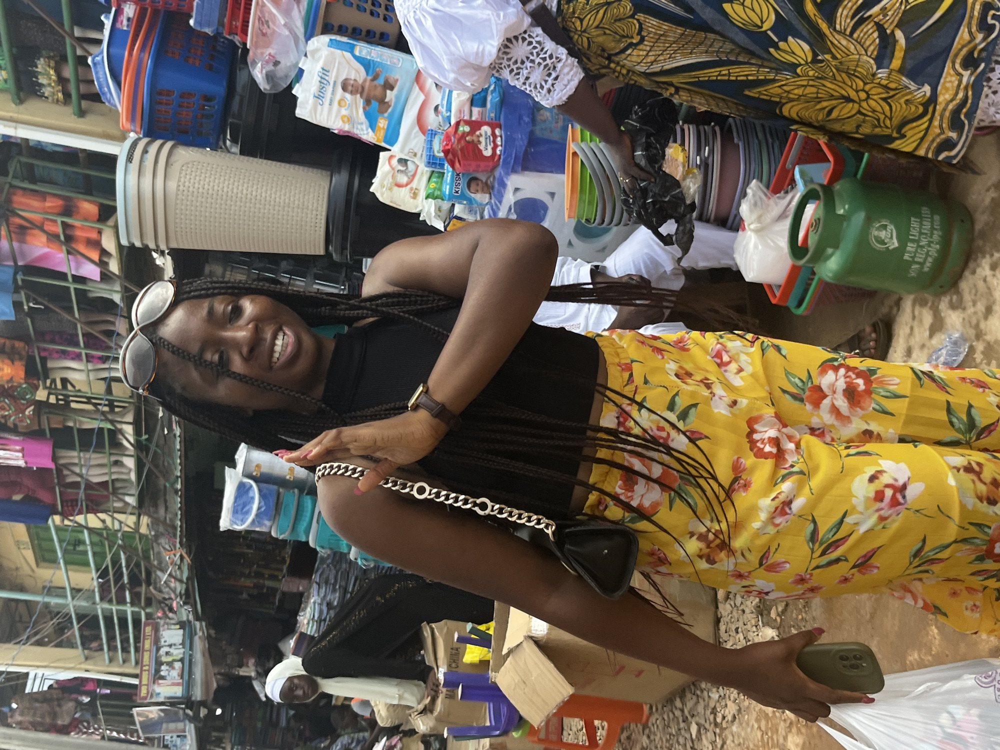 A Black woman with long braided hair and sunglasses on her head, wearing a black tank top and yellow floral pants, is standing in what appears to be a busy market in Jos, Nigeria. She is holding a white plastic bag in her left hand and a green cell phone in her right hand. She is looking to her right and smiling. In the background, there are various stalls selling goods, including stacked plastic containers, baskets, diapers, and a green gas cylinder. Another person in a white top and a patterned skirt is visible to the right of the woman. The market appears to be outdoors with a corrugated metal roof over some of the stalls.