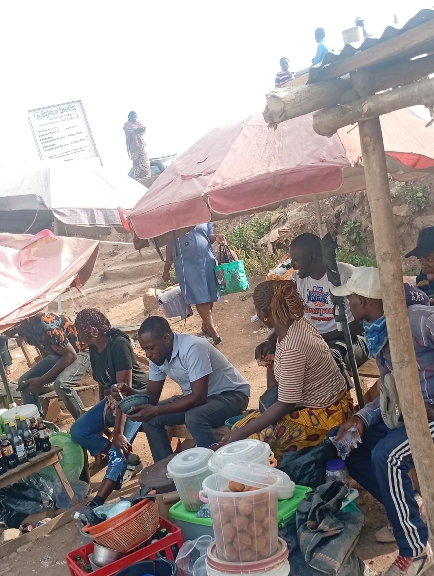 The image depicts an outdoor scene in Jos, Nigeria, showing a group of people seated under makeshift shelters and umbrellas. Some individuals are eating or drinking, while others are engaged in conversation. The scene appears to be a market or roadside food stall, with various containers and bottles visible. A woman in a light blue dress walks past, carrying a shopping bag. Above the market, a billboard reads "Rightway Assembly." In the background, two people are standing on a structure with a tin roof.
