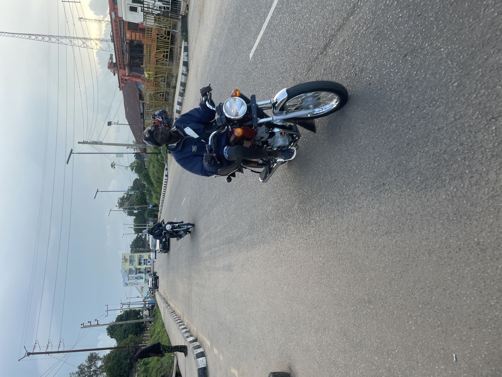 In Jos, Nigeria, a man rides a maroon motorcycle down a paved road. He is wearing a dark blue jacket, dark pants, and a beanie. The motorcycle has a large circular headlight and a chrome fender. In the background, another person is riding a motorcycle, and several cars are on the road. Buildings and utility poles line the street, with trees and vegetation visible on the sides. The sky is overcast, suggesting it might be late afternoon or early morning. The scene appears to be a typical street view in an urban or semi-urban area.