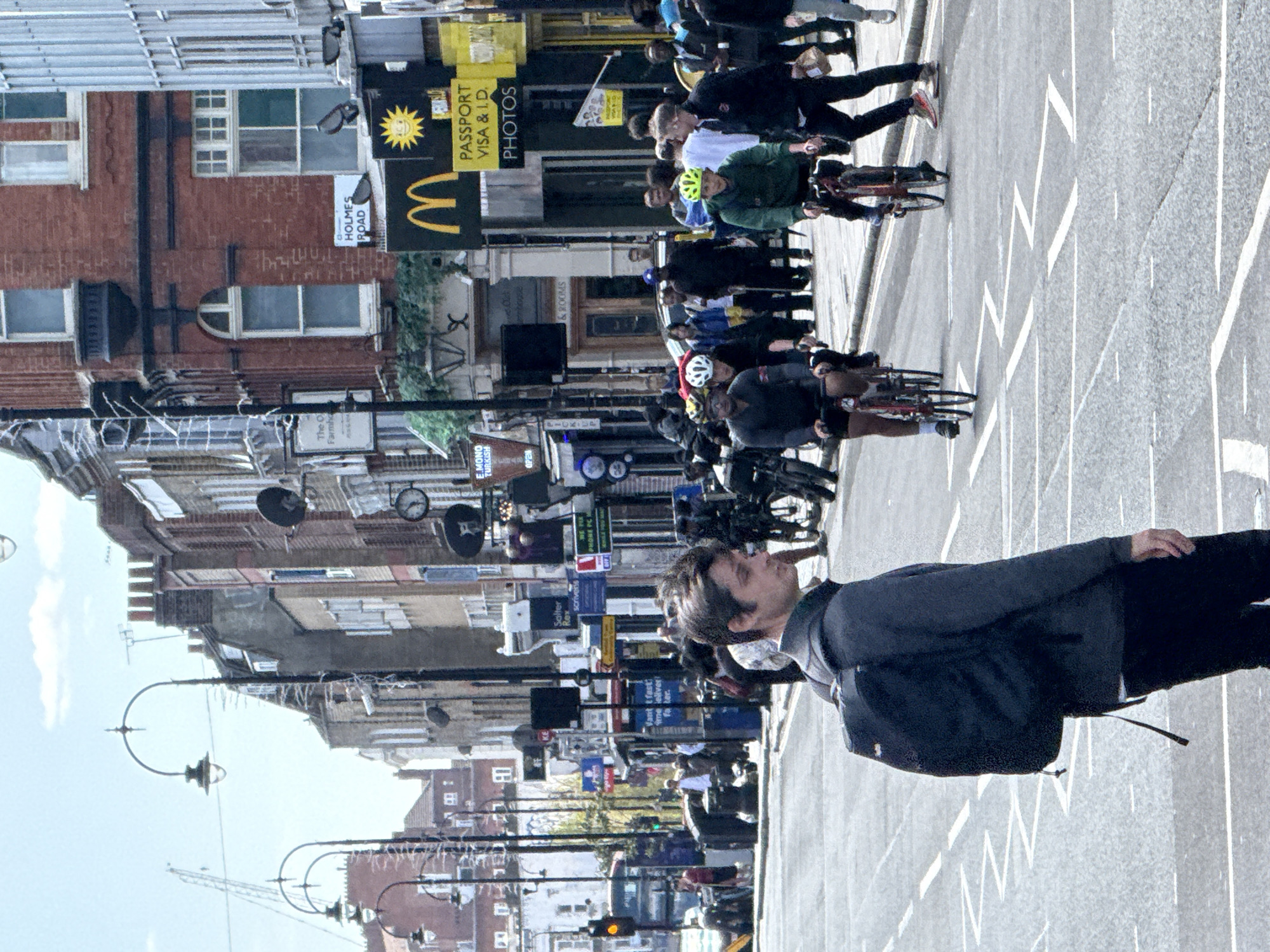 The image depicts a street scene in London, United Kingdom. A man is walking on the right side of the image with buildings behind him and to his left. On the left side of the street, there are numerous shops with awnings. Visible signs include McDonald's, Passport Visa & I.D. Photos, E. Mono Turkish Restaurant, The O Farmhca, Salter Rex, Scrivener's, Holmes Road and a sign saying "Pick up and Rooms". A large group of cyclists ride along the street. Traffic lights are present, along with street lamps. A construction crane is seen in the far distance.