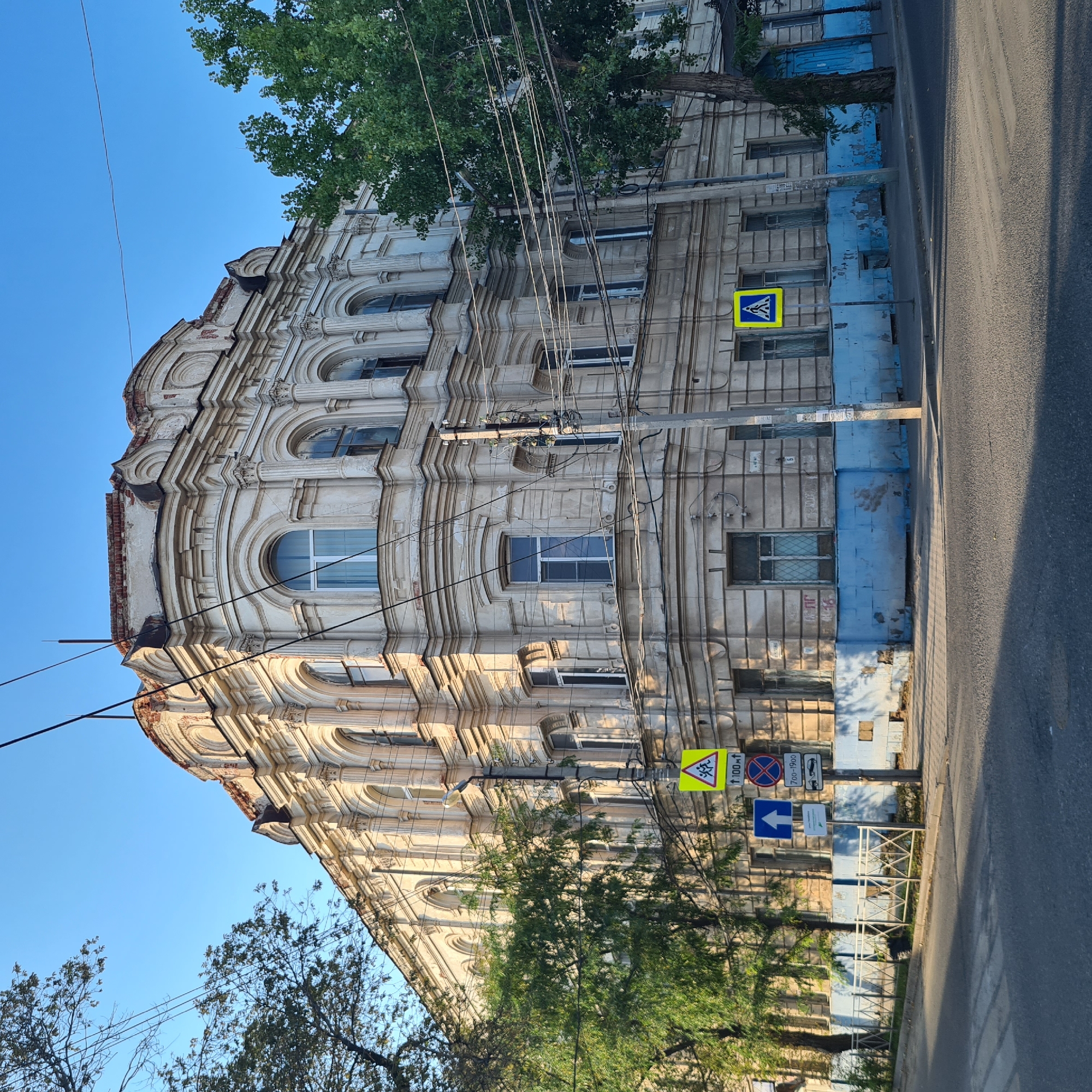 The image shows a historic, light-colored building with ornate architectural details, including arched windows and decorative moldings. The building appears to be at a street corner with trees and overhead electrical wires. Several road signs are visible: a yellow pedestrian crossing sign, a blue sign with a white arrow pointing up, a circular red and blue prohibition sign, and a sign indicating towing services between 07:00 and 19:00. The lower part of the building's facade is painted light blue, with some peeling paint and visible brickwork in places. A white metal fence runs along the sidewalk. The street in the foreground is asphalt with a marked pedestrian crossing. The sky is clear and blue. The architectural style and street signage suggest the location is in Astrakhan, Russia.