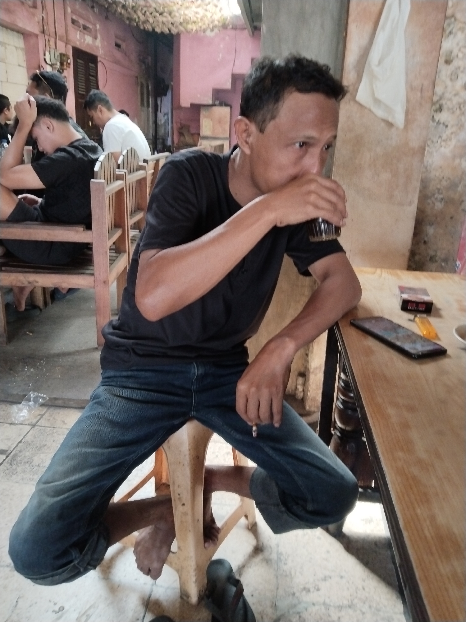 The image depicts an outdoor or semi-outdoor scene, possibly a cafe or a roadside stall, in Kecamatan Pati, Indonesia. In the foreground, a man is seated on a light-colored plastic stool, wearing a black t-shirt and blue jeans. He is holding a glass in his right hand and appears to be drinking from it. His legs are splayed apart, and his bare feet are visible.
To the right of the man, a wooden table is visible. On the table are several objects: a smartphone, a pack of cigarettes, a lighter, and a cup. In the background, several other individuals are seated at wooden tables and chairs. One person in the mid-ground, also wearing a black t-shirt, has their head resting on their hand, looking down. Another person, wearing a white t-shirt, is also seated and looking downwards. The environment appears somewhat aged, with pink-colored walls and a woven ceiling. The ground is tiled. The overall impression is of a casual gathering or a moment of rest.