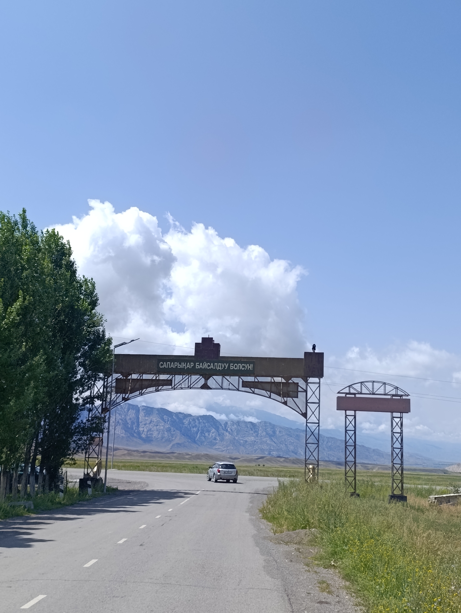 The image depicts a road leading towards an archway with text in Cyrillic script. The archway is constructed of metal and supports a sign with the words "САПАРЫНАР БАЙСАЛДУУ БОЛСУН!". To the left of the archway is a tall, leafy green tree and a wooden fence. A silver car is visible driving away from the camera and under the archway. Beyond the archway, a vast, flat landscape stretches out, with mountains visible in the distance. The sky is a clear blue with scattered white clouds. The overall scene suggests a rural or semi-rural setting, possibly an entrance to a town or region. Based on the Cyrillic text and mountainous backdrop, the location is identified as Emgekchil, Kyrgyzstan.