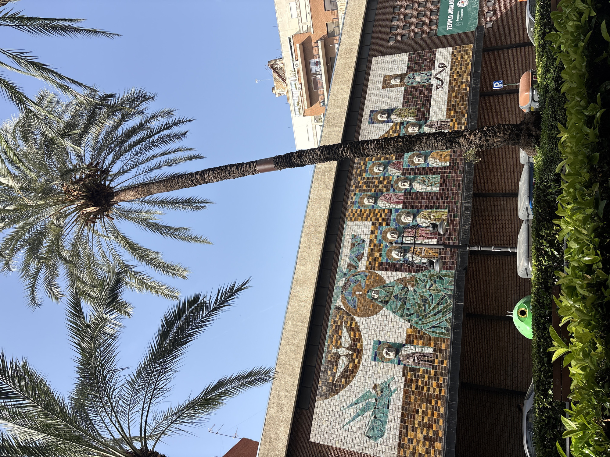 The image shows a street scene in Valencia, Spain, with a large mosaic mural on the facade of a building. The mural depicts religious figures and symbols in a stylized, colorful manner. In the foreground, there are palm trees, lush green bushes, and a dark wooden bench. Behind the bushes, several trash receptacles are visible, one of which is a distinctive bright green, round container with a large eye-like opening. A light-colored car and a gray trash bin are also present. A street lamp stands near the mosaic. Further in the background, the building facade continues with a brick pattern and a sign that reads "TEMPLO JUBILAR". Other buildings with balconies and windows are visible above the mosaic. The sky is clear and blue. The overall impression is a sunny day in an urban environment with a significant piece of public art.