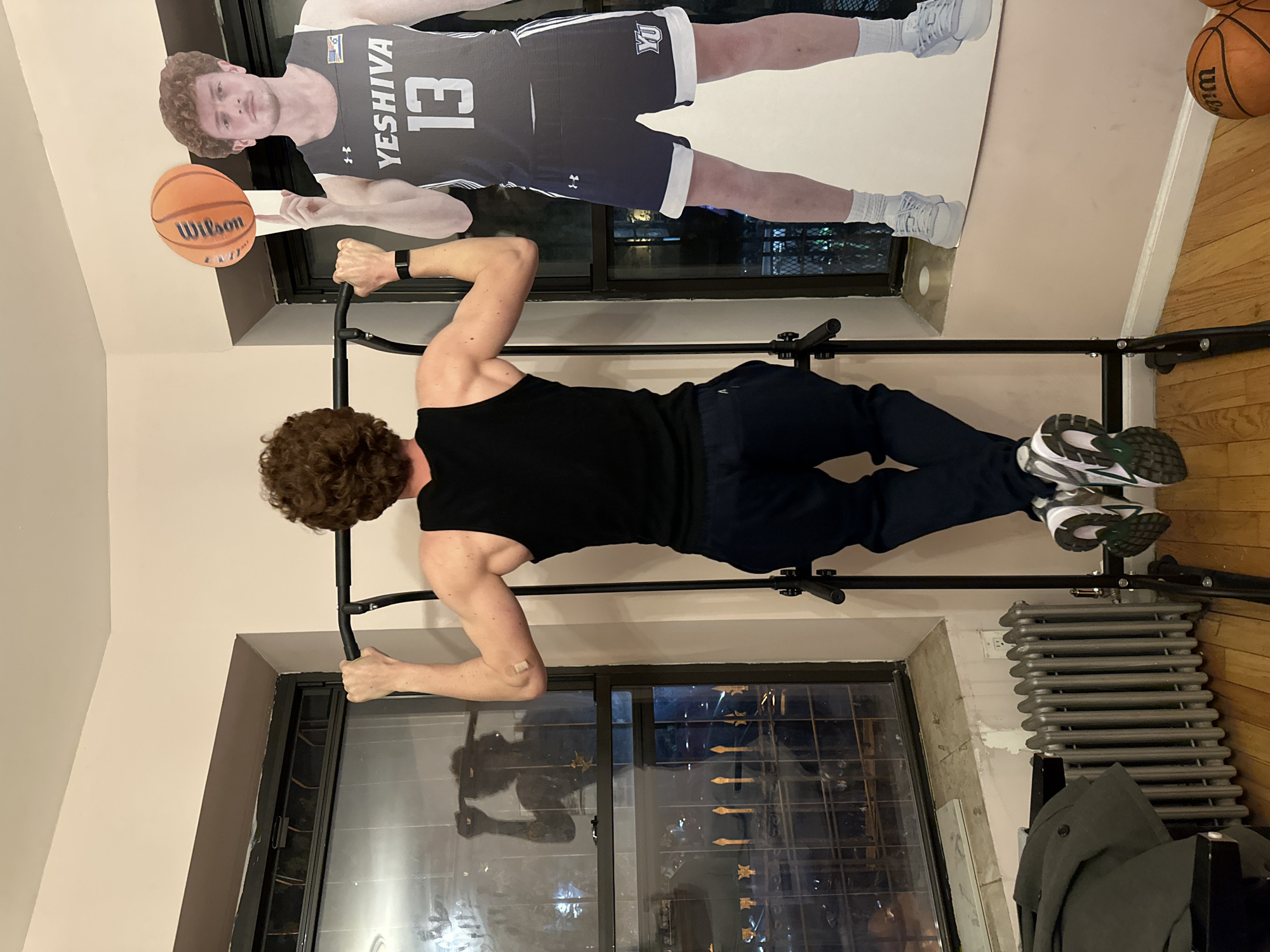 This indoor image, rotated 90 degrees counter-clockwise, depicts a man exercising on a pull-up bar, with a life-sized basketball player cutout nearby.
The subject is a man with curly brown hair, seen from behind, wearing a black tank top, dark athletic pants, and white and green athletic shoes. He has a black smart-watch-like band on his left wrist and a small bandage on his left elbow. He is actively performing a pull-up, gripping the bar with both hands, his arms bent and body elevated. The pull-up bar is mounted above a window.
To the man's right, a large cardboard cutout of a male basketball player stands against the wall. The cutout wears a grey basketball jersey featuring "YESHIVA" and the number "13", with a small "Under Armour" logo. His shorts also display "YU". A "WILSON" branded basketball is positioned to appear as if held by the cutout's hand.
Behind the man, through the window, reflections of city lights and buildings suggest an urban environment at night or evening. Below the window, a grey cast-iron radiator is visible. The flooring is wood. Another basketball is partially seen on the floor near the radiator, and a dark jacket or coat is draped over an object below the radiator. The setting appears to be an apartment in an urban area, aligning with the New York, USA context implied by "Yeshiva University".