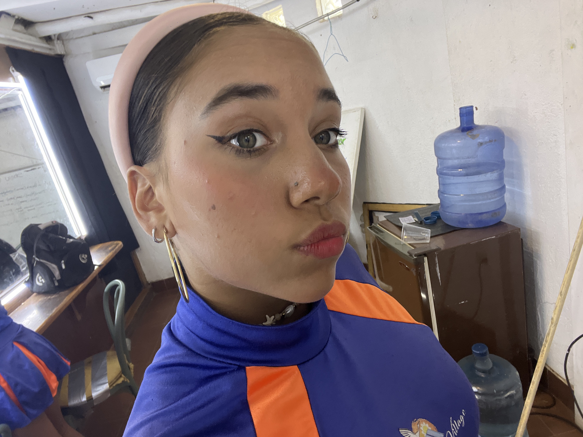 A close-up shot of a young woman with olive skin, dark eyebrows, and green eyes. She has a piercing on the left side of her nose and is wearing a light pink headband. She is wearing a blue and orange athletic top and teardrop earrings. She is looking at the camera with her lips pursed. In the background, there is a desk with items on it and a large bottle of water on top of a brown cabinet. There's also a wooden pole leaning against the white wall. The location is likely indoors, but it is not possible to determine if it is in the city of Porlamar, Venezuela, based on the visual information provided.