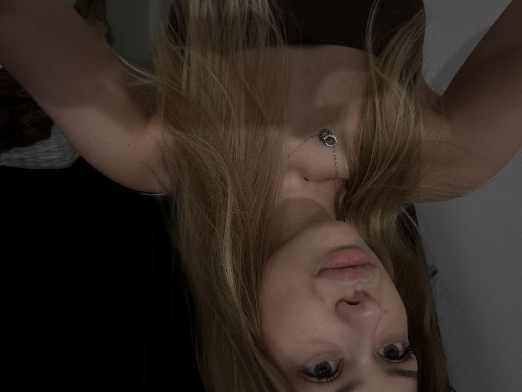 This is a close-up selfie of a young Caucasian woman with long, blonde hair. She is wearing a dark brown strapless top and a silver necklace with two interlocking rings. Her lips are full and painted with a pinkish-nude lipstick. The background is dark and out of focus, with hints of a grey wall on the left and a dark surface on the right. There is no discernible connection to Đà Nẵng, Vietnam.