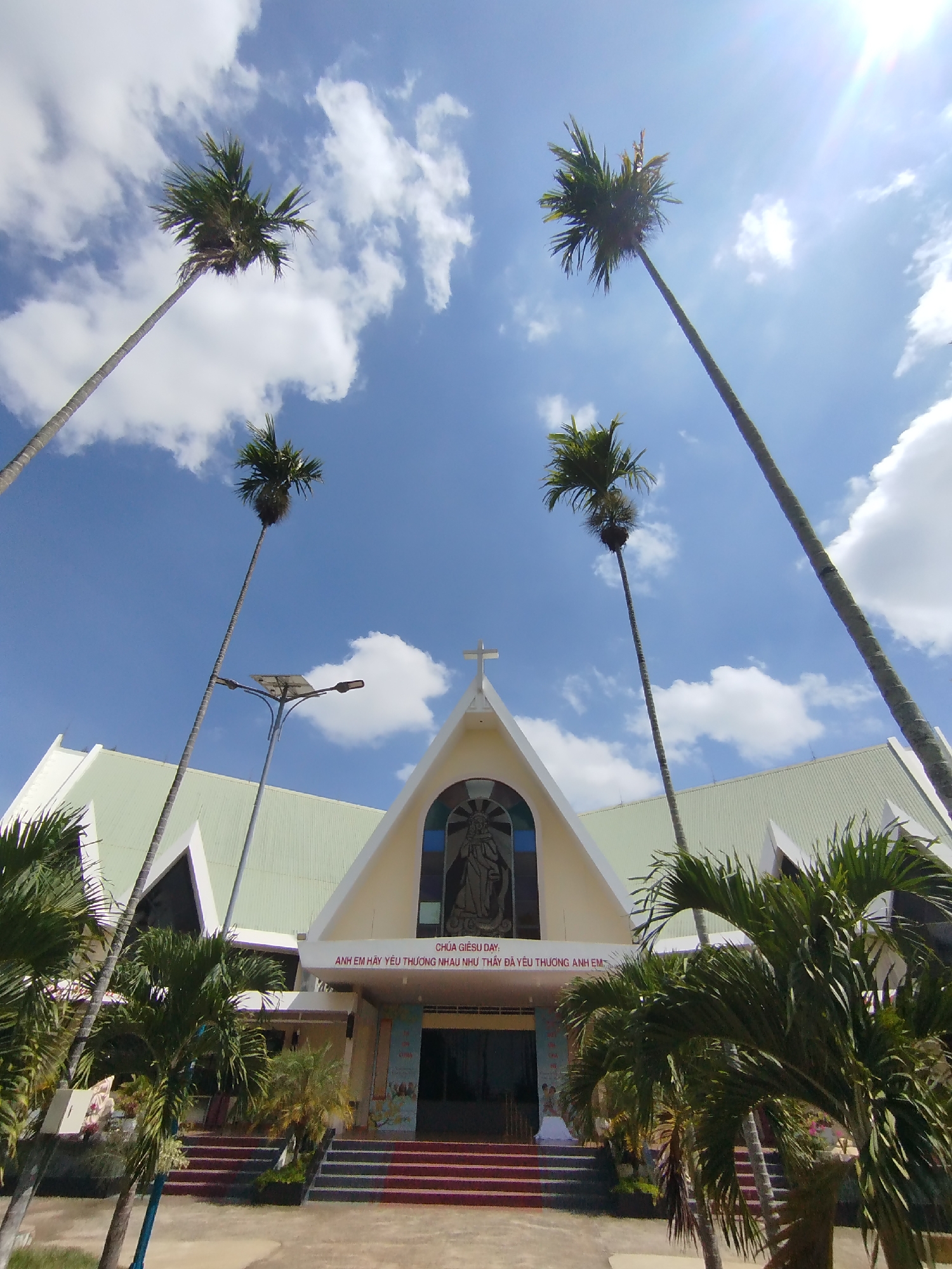 The image shows a low-angle view of a church in Pleiku Ward, Vietnam. The church has a simple, modern design with a gabled roof and a cross at its apex. In front of the church are several tall, slender palm trees, reaching up into the blue sky dotted with white clouds. Smaller, bushier palm trees are also present, framing the entrance and adding a tropical feel. A light pole stands to the left of the church. A sign above the church entrance displays text in Vietnamese, "CHÚA GIÊSU DẠY: ANH EM HÃY YÊU THƯƠNG NHAU NHƯ THẦY ĐÃ YÊU THƯƠNG ANH EM," suggesting a religious message. The scene is bathed in sunlight, giving it a bright and cheerful atmosphere.