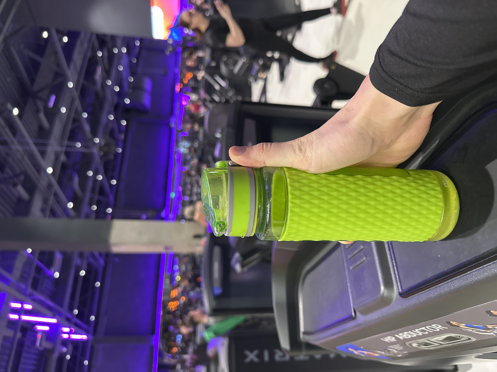 A person is holding a light green water bottle in what appears to be a fitness center. The water bottle is textured and partially open, with a visible water droplet. The person is seated on or near a "HIP ABDUCTOR" machine from the brand "MATRIX." Other individuals are exercising in the background. The background includes stage lighting and structural elements of a large building. The location is likely Murino, Russia, given the prompt's instructions.