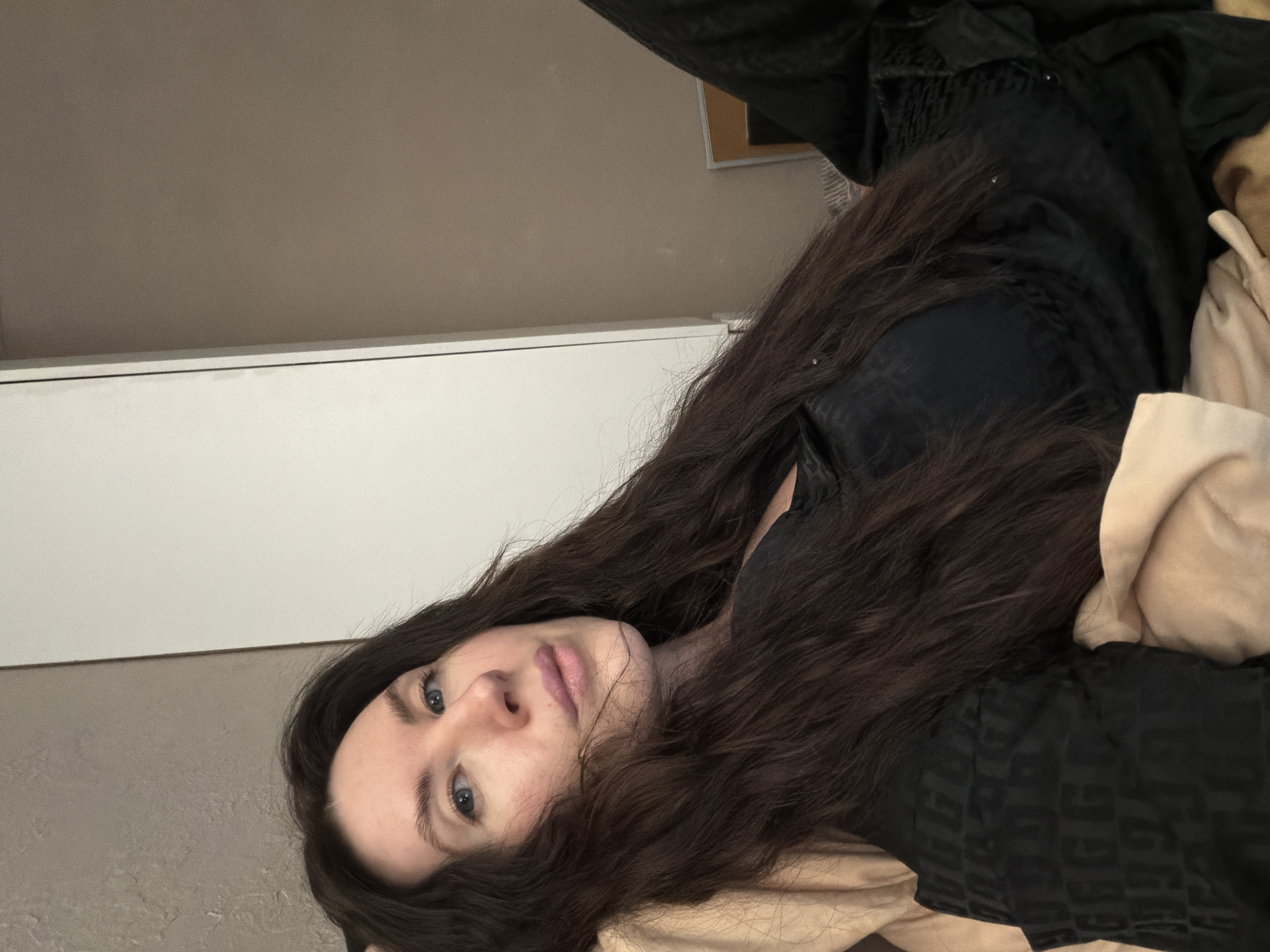 A single individual, an adult female, is depicted in a medium close-up, lying indoors on a light-colored pillow or bedding. Her head is angled slightly, and she looks directly towards the camera. She has long, dark brown hair, fair skin, blue eyes, and light pink lips. She is wearing a dark garment; the visible left shoulder area features a subtle, repeating 'G' pattern, while the right side of the garment is a plain dark fabric, possibly satin, with some gathered detail. Behind the individual, a plain light brown wall is visible on the right, and a white, tall, vertical furniture piece, likely a wardrobe or cabinet, is positioned on the left. In the lower right portion of the background, a partially obscured framed object with a light brown border is visible, resting against the wall above a section of striped material, possibly additional bedding. The specific location within Perm, Russia, cannot be determined from the visual information provided.