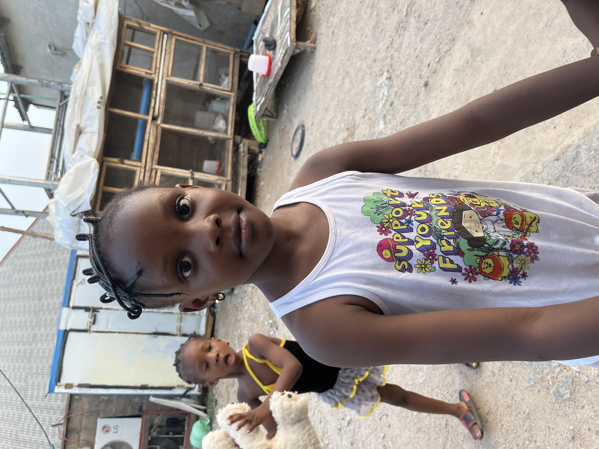 The image depicts two young girls in Madalla, Nigeria. The foreground features a girl with braids, wearing a white tank top with a colorful cartoon print. Behind her, another girl is visible, holding a white stuffed animal and wearing a black and yellow dress. The setting appears to be outdoors, possibly in a residential area, with structures visible in the background. These structures include what seems to be some sort of building, air-conditioning outdoor unit, and wooden cages that could be for animals. The ground appears to be dirt or gravel.