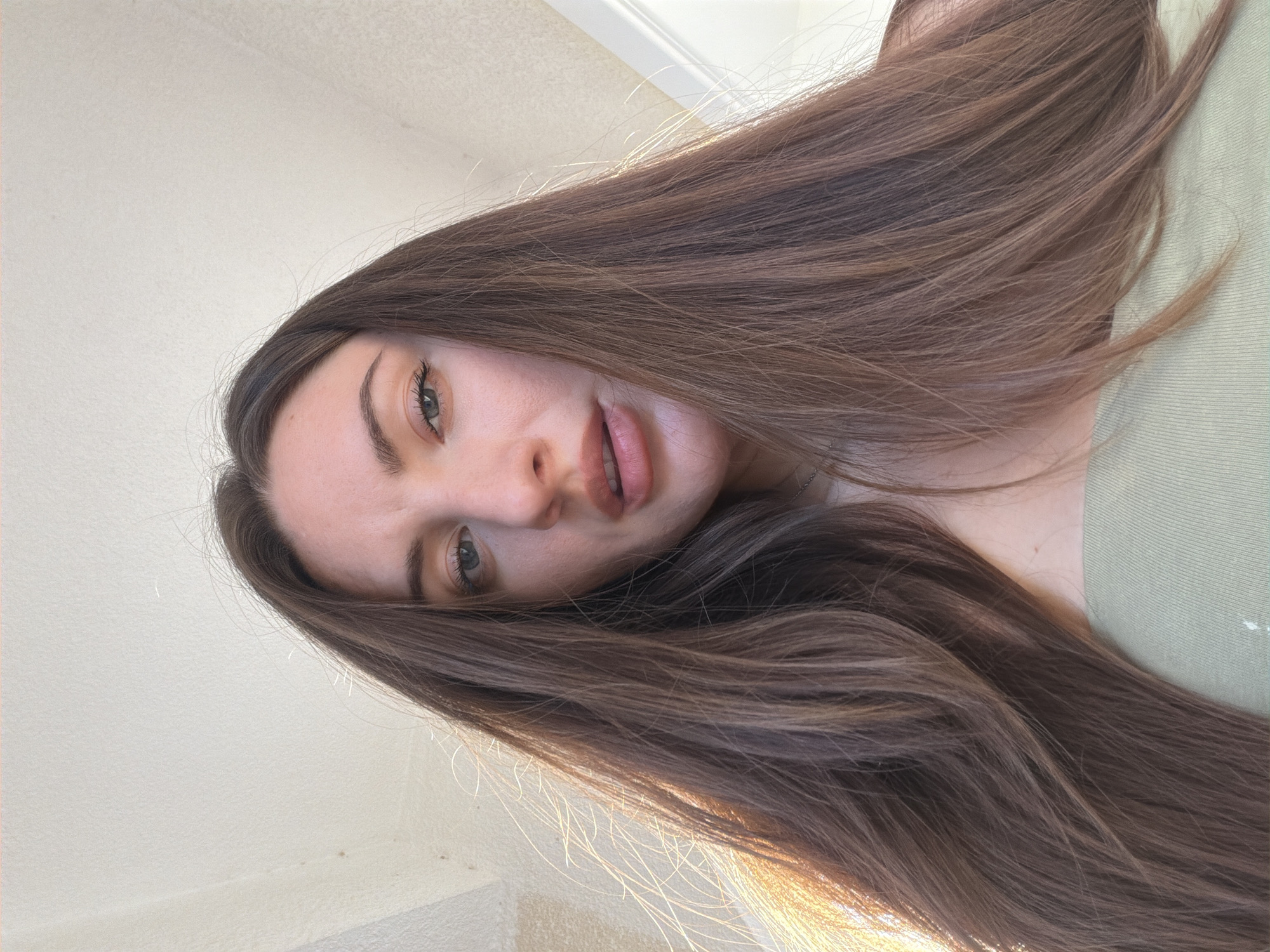 The image shows a low-angle, close-up shot of a young woman with long, brown hair. She is looking directly at the camera with a neutral expression. Her hair is spread out around her face and shoulders. She is wearing a light green, off-the-shoulder top. The background appears to be an indoor setting with a textured wall and ceiling, suggesting a room with diffused natural light. There are no discernible landmarks or elements that would place the image specifically in Sharm El Sheikh, Egypt.