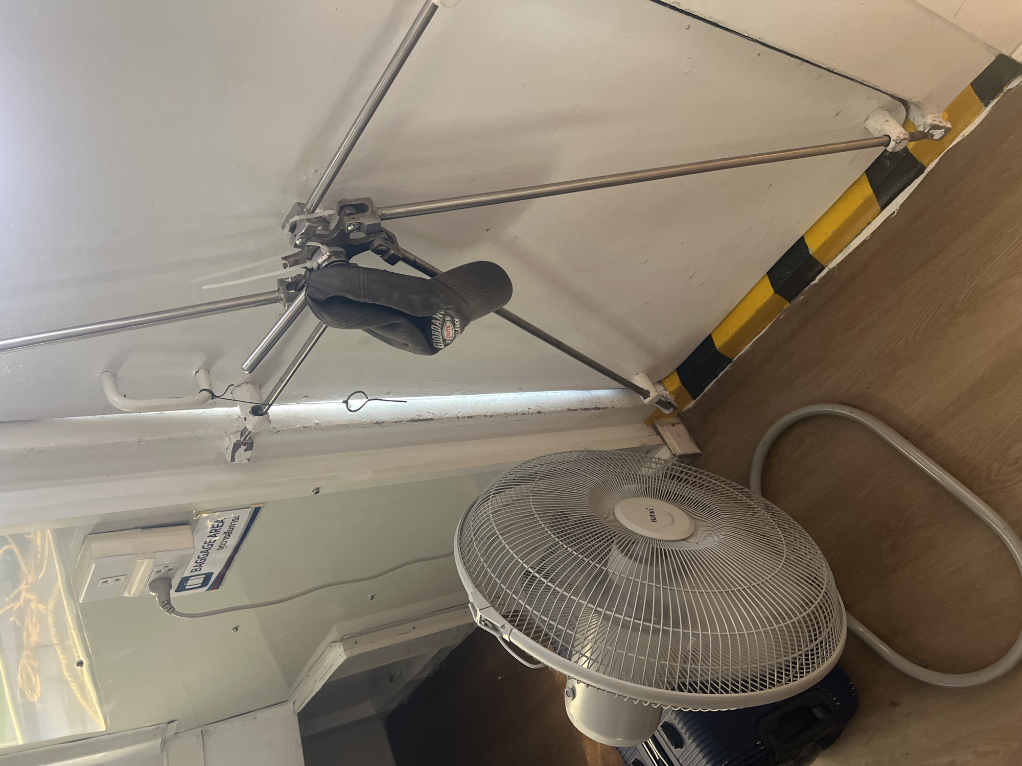 The image shows an interior scene, possibly on a ferry or boat given the metal structure and the proximity to what appears to be a door or bulkhead. In the foreground, a white oscillating fan with the brand name "Hatari" is positioned on a wooden floor. To the left of the fan, a portion of a dark blue suitcase is visible. A power outlet with a plugged-in cord is mounted on a white wall to the left, above a sign indicating "BAGGAGE AREA" in English and Thai. A black cap with the word "GIORDANO" is hanging from a metallic structure composed of several poles that extend diagonally across the frame, attached to the wall by various clamps and zip ties. The wall itself is painted white, with a black and yellow striped bumper guard along the bottom. The overall environment appears utilitarian and functional. The location in ตำบลเกาะพะงัน, Thailand, suggests a tropical setting, potentially a mode of transportation to or from the island.