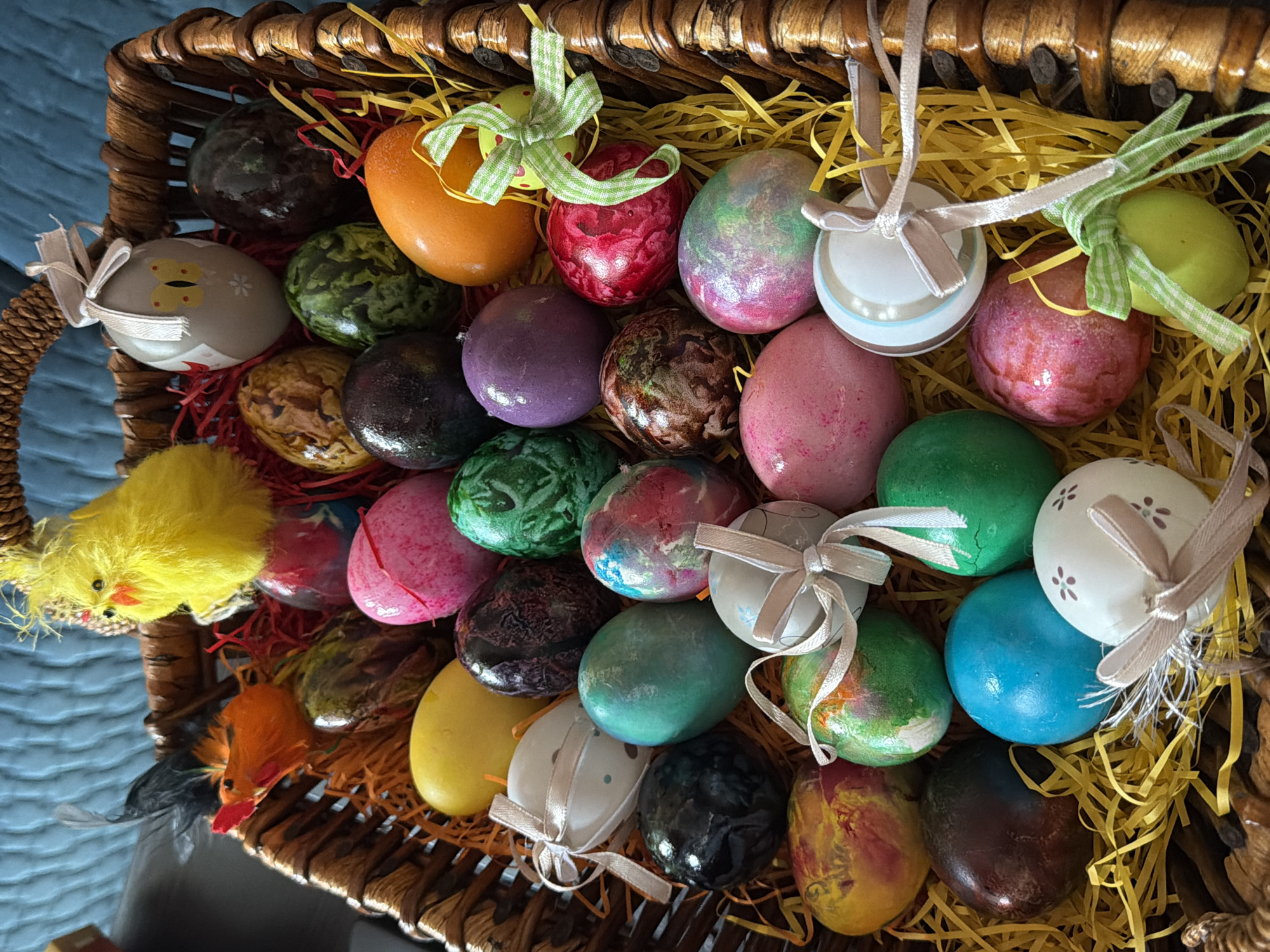 A high-angle, close-up shot shows a wicker basket filled with Easter eggs. The eggs are multi-colored and decorated with ribbons and patterns. Some decorative chicks are visible near the left edge of the basket. The basket is filled with yellow straw. The image appears to be taken indoors, possibly in a home, though specific location information such as Sofia, Bulgaria, cannot be confirmed visually.